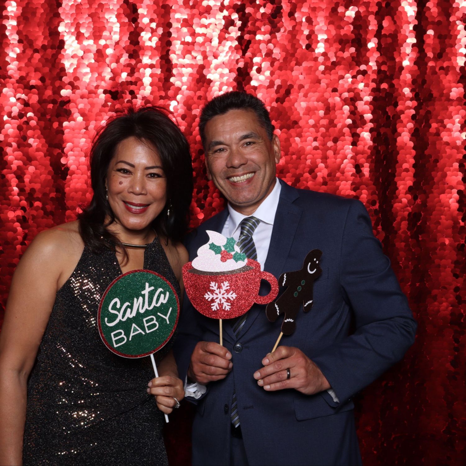 Couple posing with Santa Baby and Christmas hot cocoa photo booth props in front of red sequin backdrop at the Gensler San Francisco party