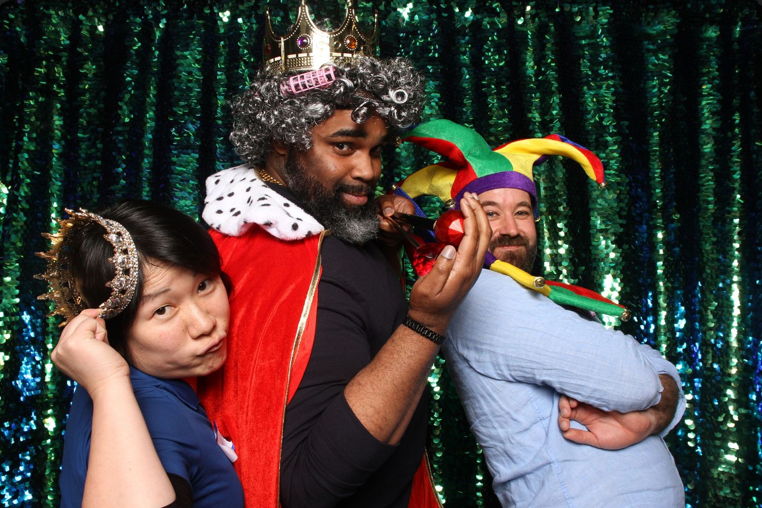 Three people in festive costumes including king and jester hats posing at photo booth with green sequin backdrop.