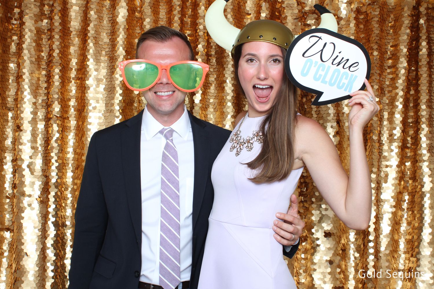 Couple posing in photo booth with gold sequin backdrop, wearing fun props including oversized glasses and viking hat.