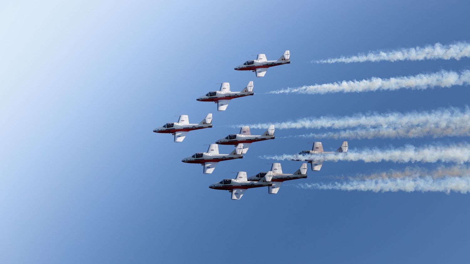 Seven military jets fly in tight formation against a clear blue sky, leaving white smoke trails at an airshow.