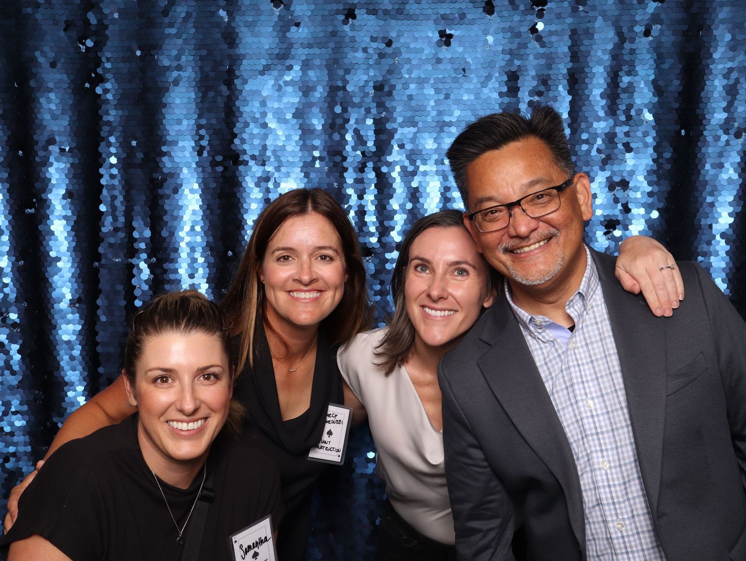 Four smiling friends posing together in front of a blue sequin backdrop at a festive event.