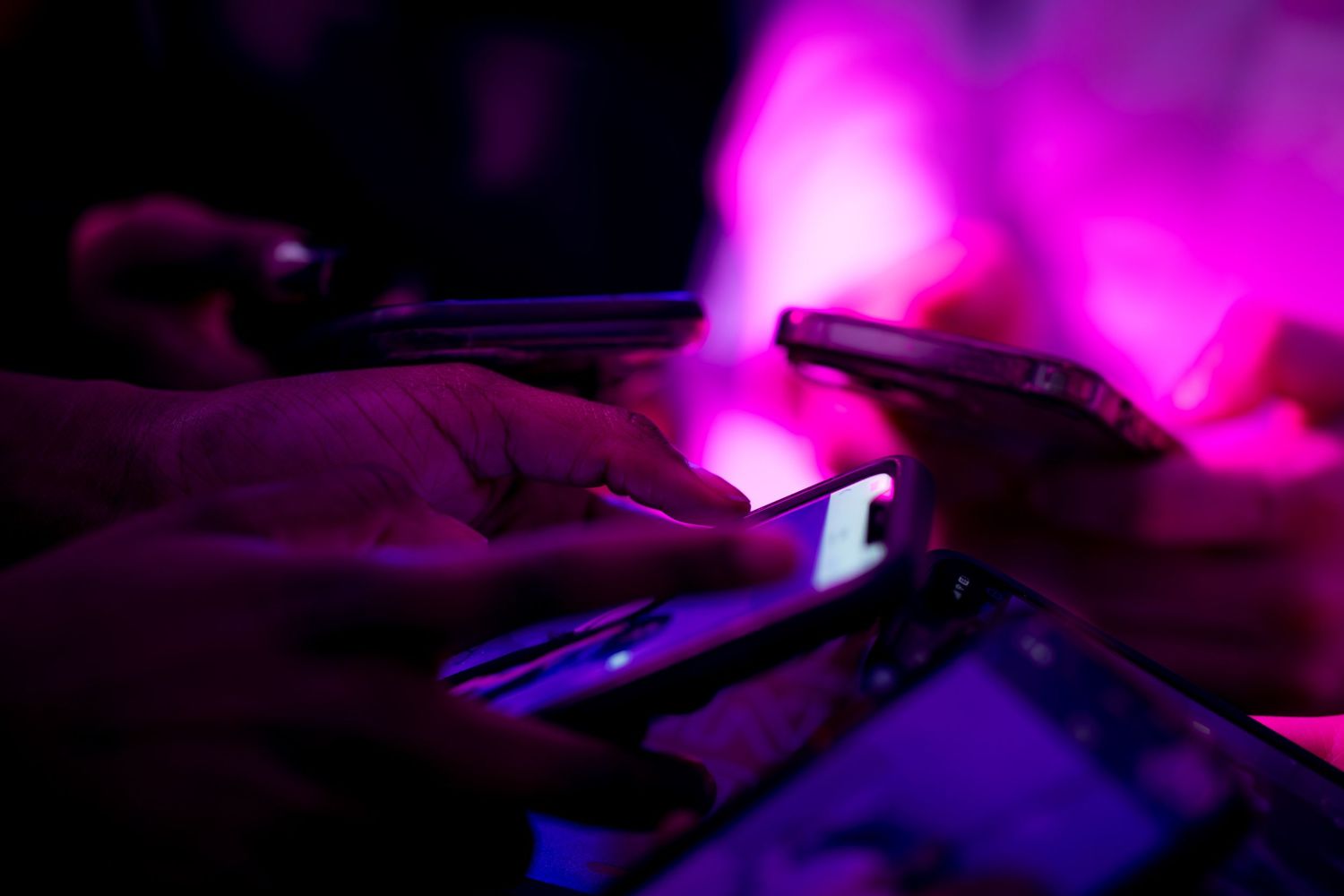 Hands using smartphones under vibrant pink and purple neon lights at a nightclub event.