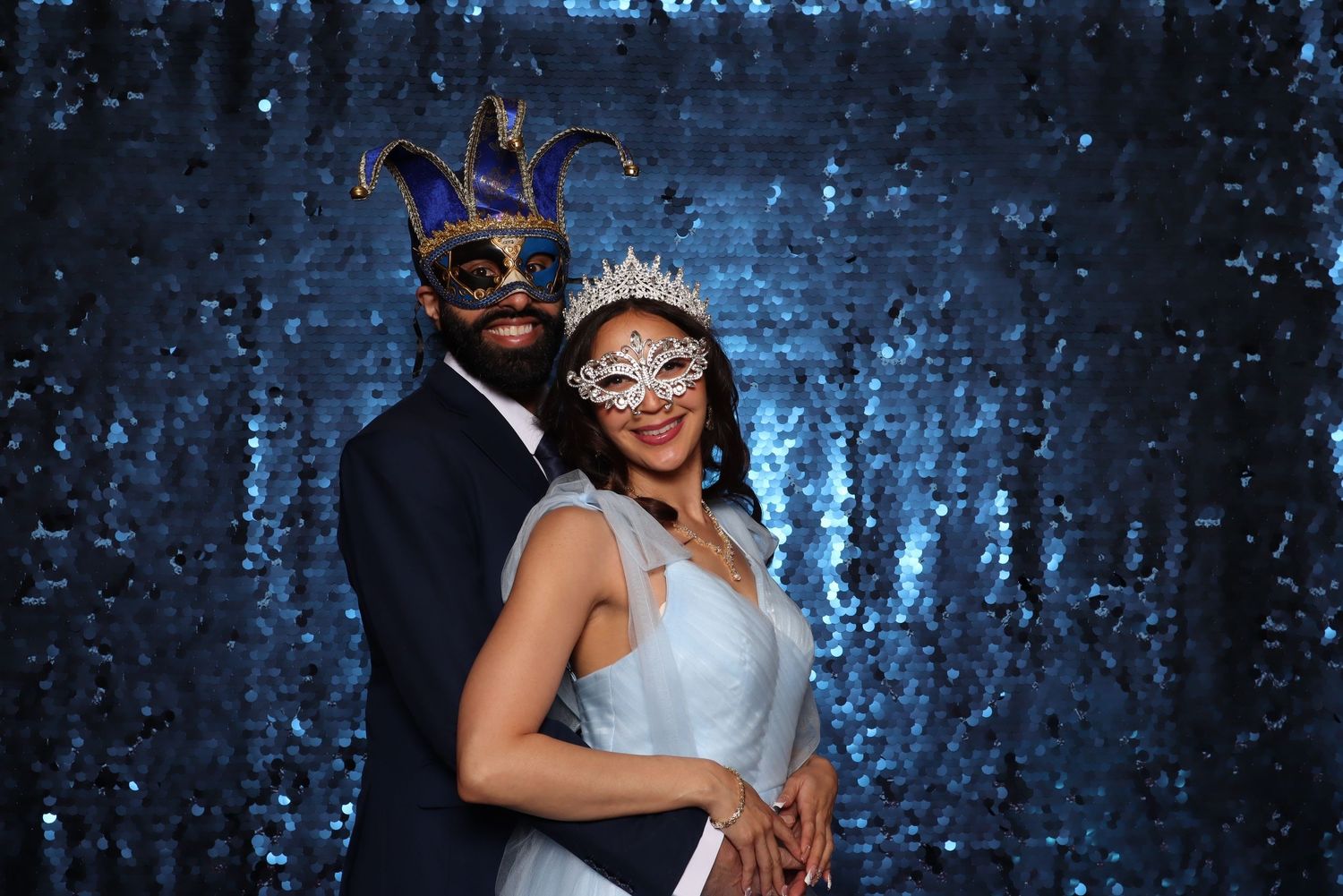 Couple in masquerade masks posing in front of blue sequin backdrop at formal event.