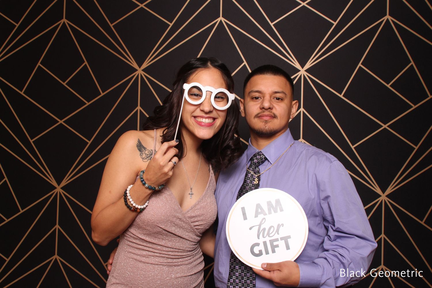 Couple posing with fun photo booth props including oversized glasses and an I Am Her Gift sign.