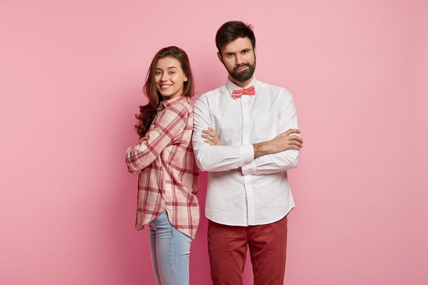 Smiling woman and serious man with bow tie standing back-to-back with arms crossed on pink background.