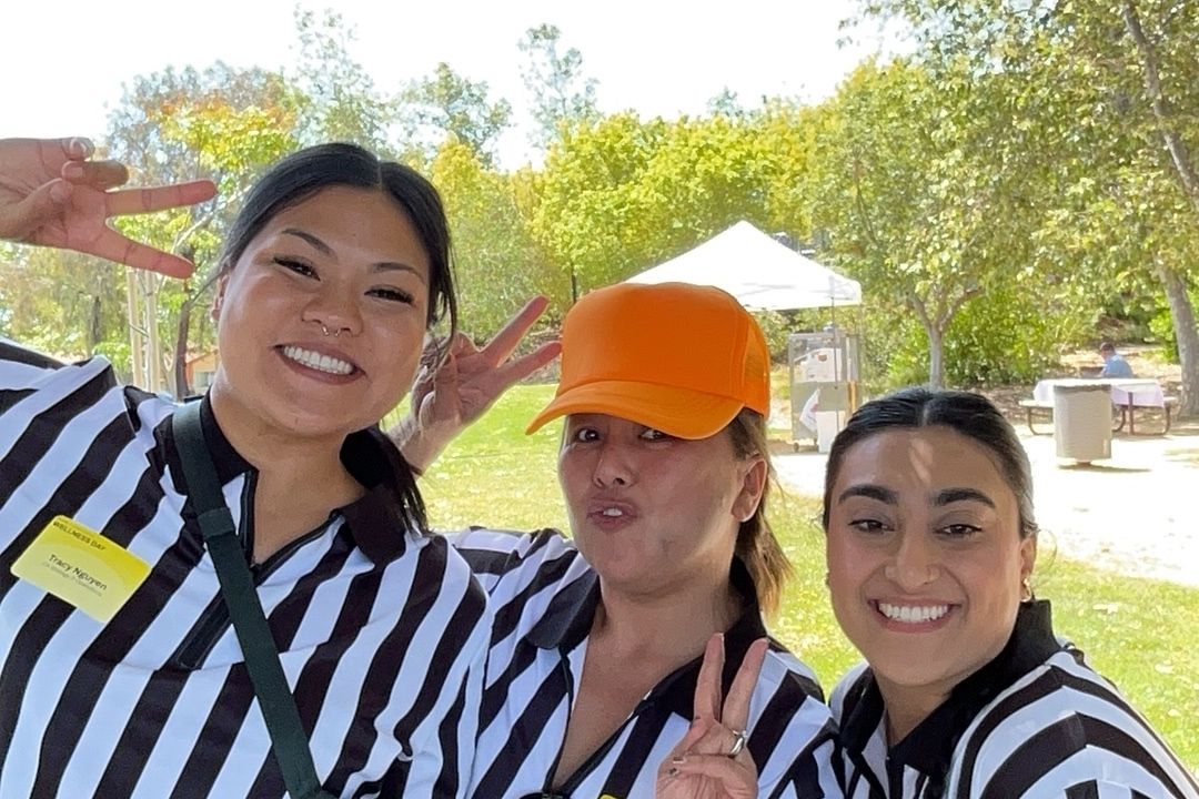Three women in referee uniforms smiling outdoors, one wearing an orange cap, posing together at a sunny park event.