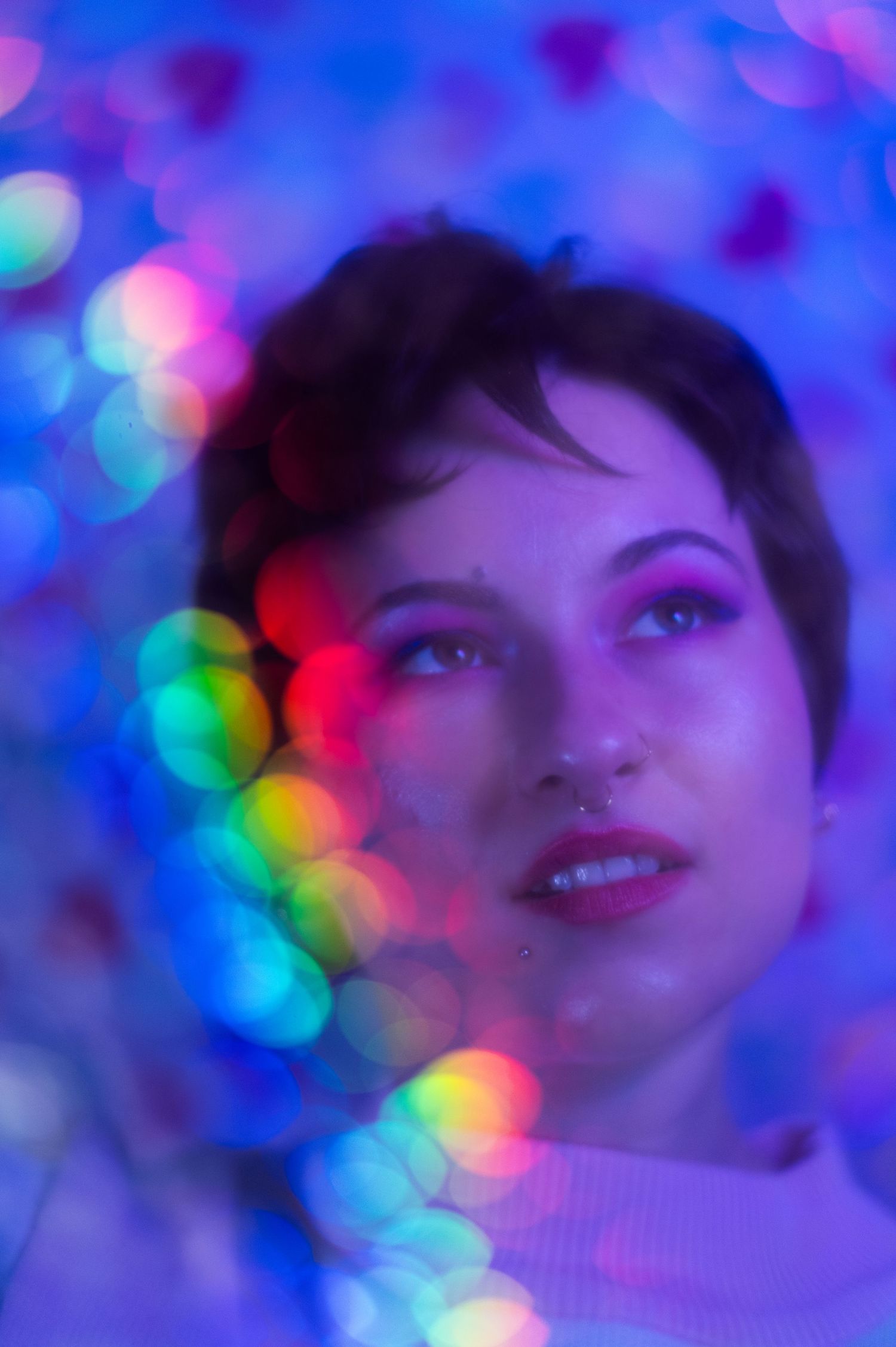 A non-binary person with pink eyeshadow and a nose ring gazes upward, surrounded by colourful bokeh lights in blue and rainbow hues.