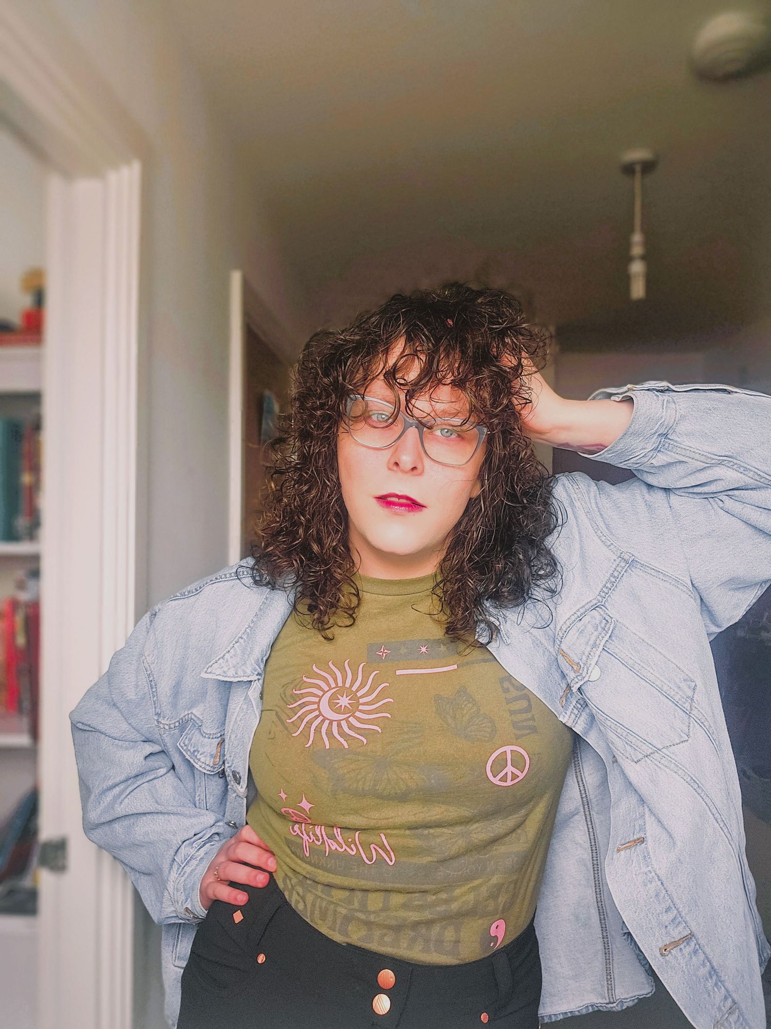 Curly-haired woman in glasses, denim jacket, and olive graphic tee poses confidently indoors.