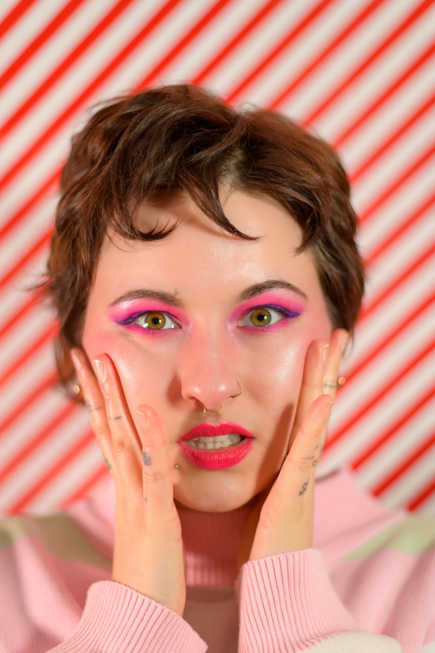 Young non-binary person with pink eyeshadow, red lips, and short brown hair poses against red and white striped background.