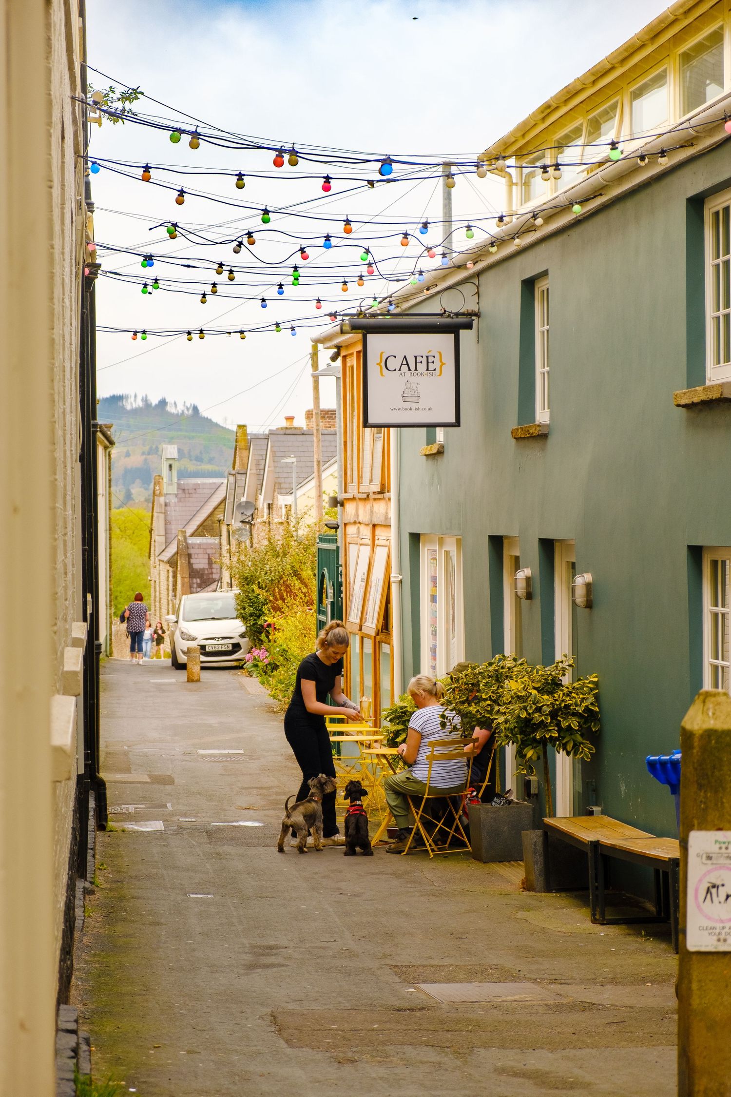 Charming narrow village street with colourful string lights, a cafe, and people relaxing outside on a cloudy day.