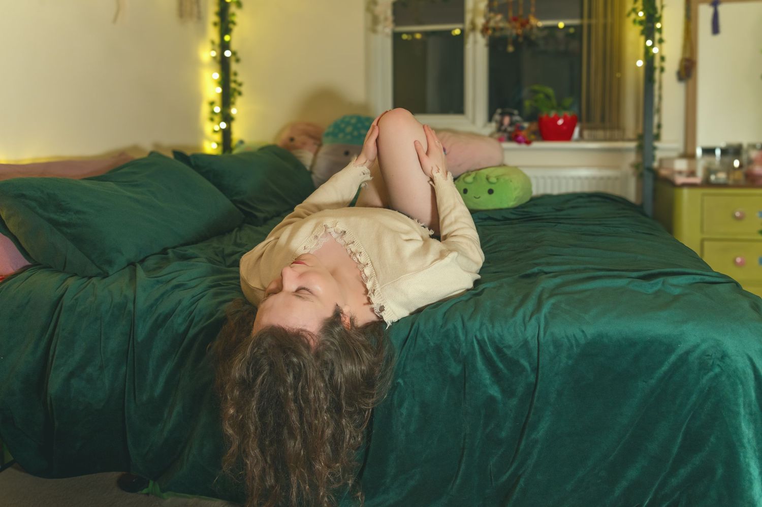 A young woman with curly brown hair relaxes on a green velvet bed with matching pillows in a cosy bedroom with fairy lights.