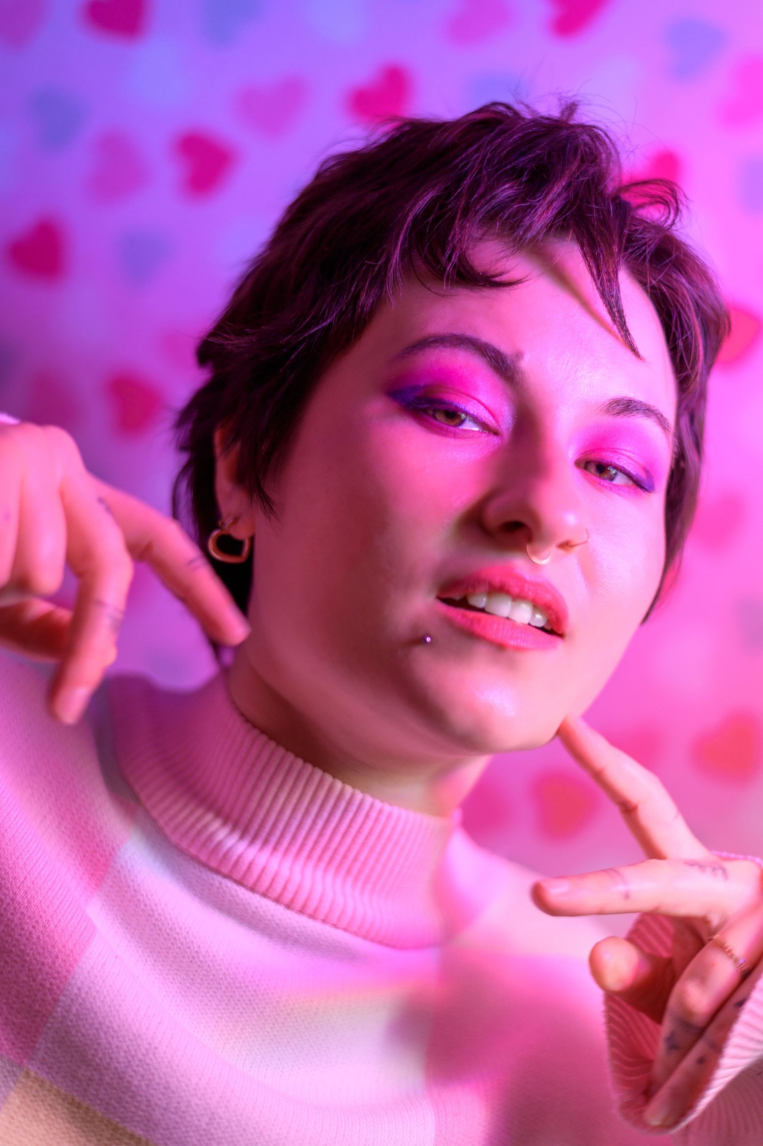 Young non-binary person with short hair and bold pink eyeshadow poses playfully in pink neon light with heart backdrop.