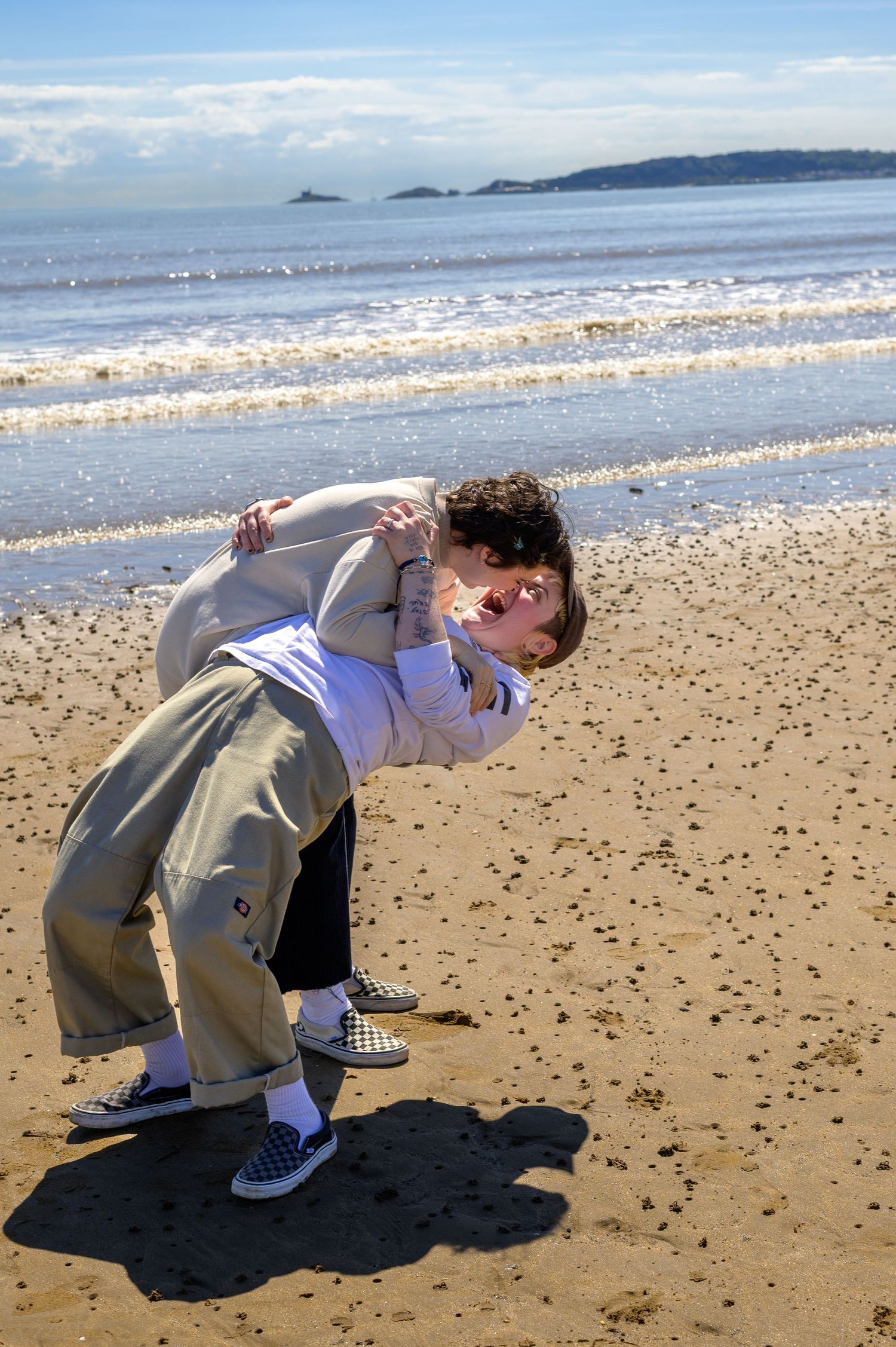Two people playfully embrace on a sandy beach with ocean waves and blue sky in the background.