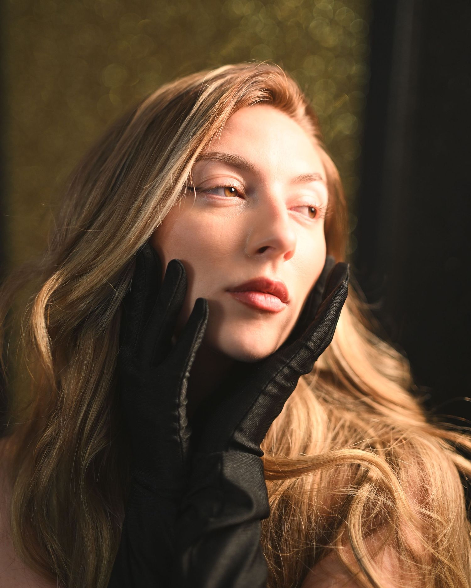 A young woman with long blonde hair wearing black gloves poses elegantly in warm golden light.