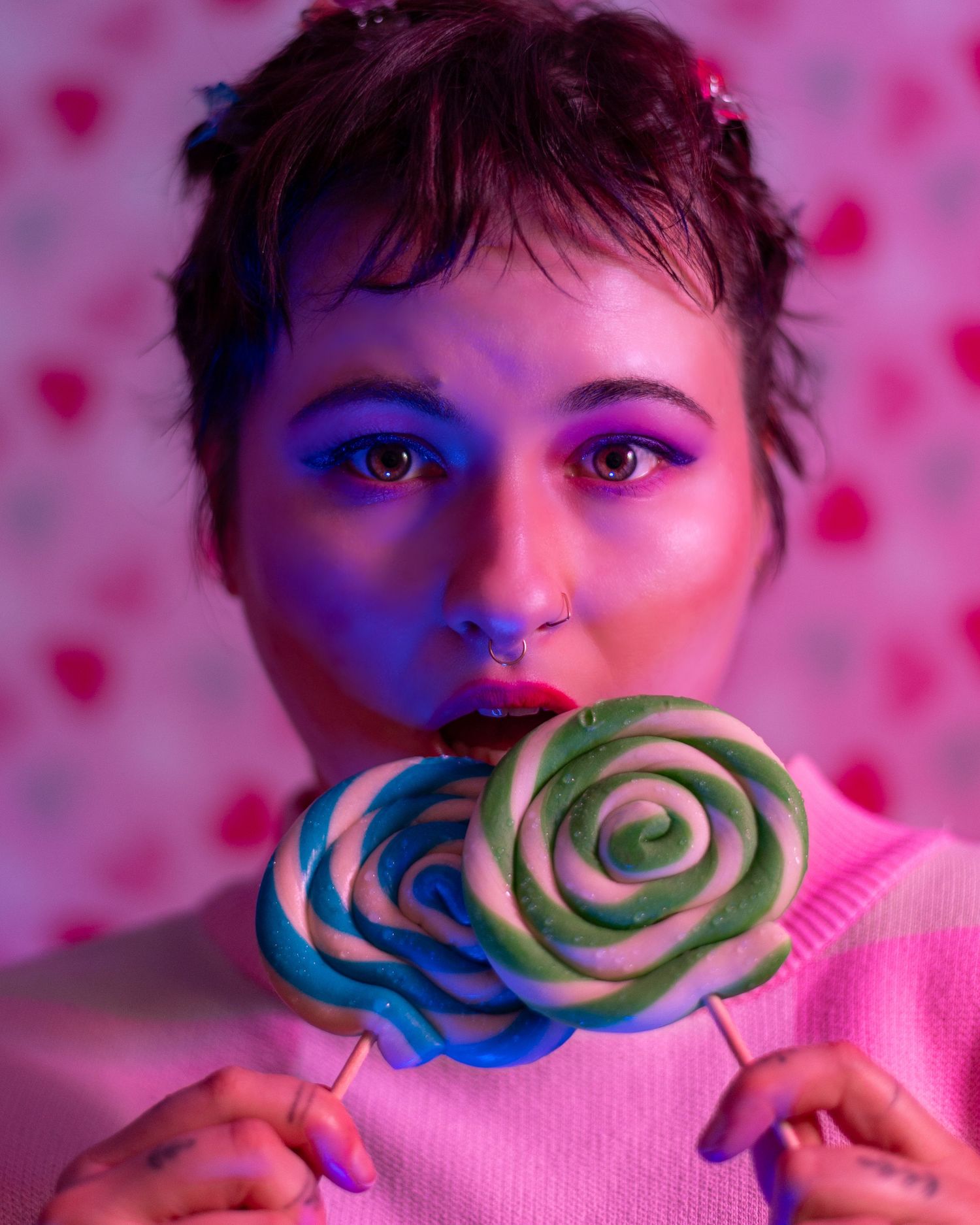 A Non-binary person with colourful purple makeup holding two swirled lollipops under neon pink and blue lighting.