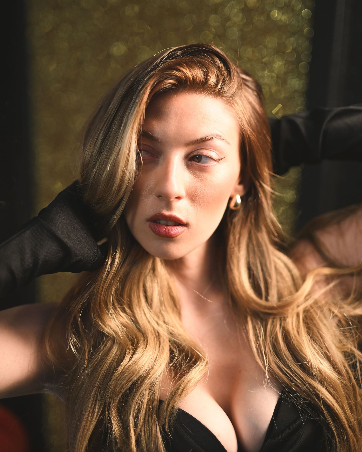 A glamorous woman with long wavy blonde hair wearing black gloves and a dark top poses against a gold backdrop.