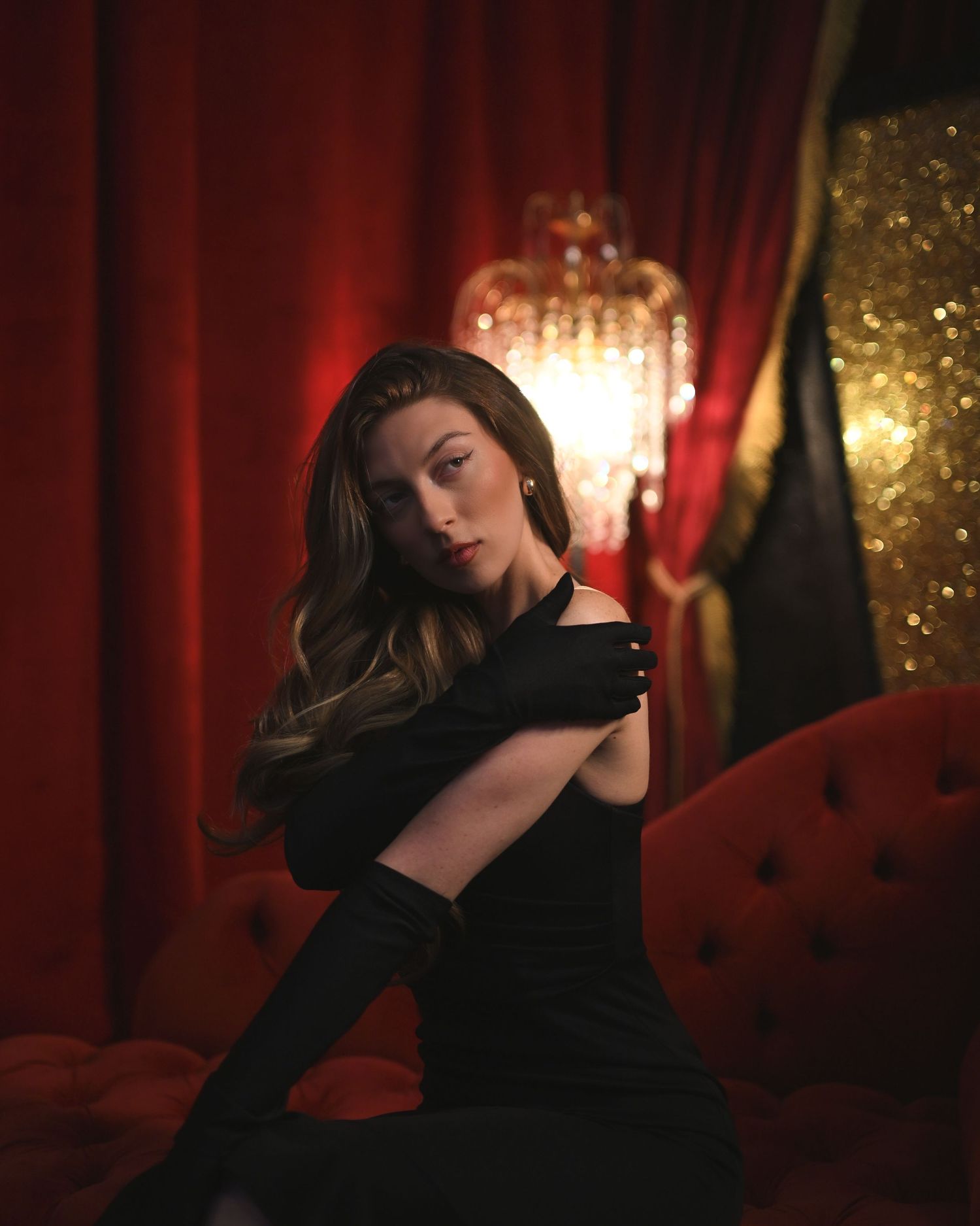 A woman in a black off-shoulder dress and long gloves poses elegantly on a red velvet sofa with a chandelier backdrop.
