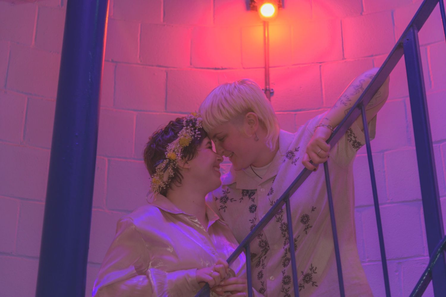 Two people in white outfits share a romantic moment on a staircase under a pink and purple light.