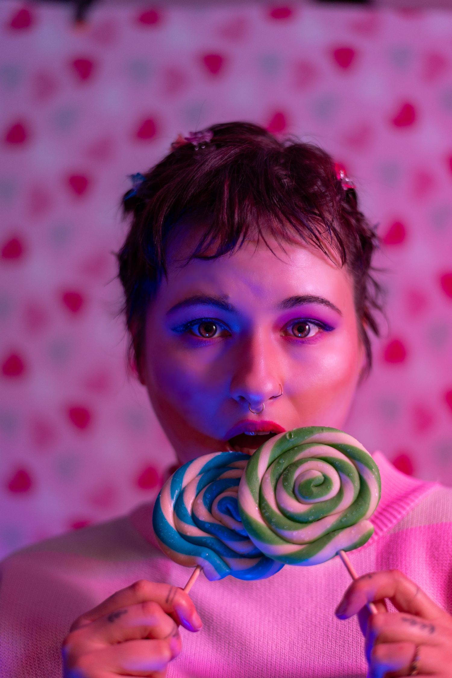 Young non-binary person with colourful makeup holding two swirled lollipops under pink and blue neon lights.