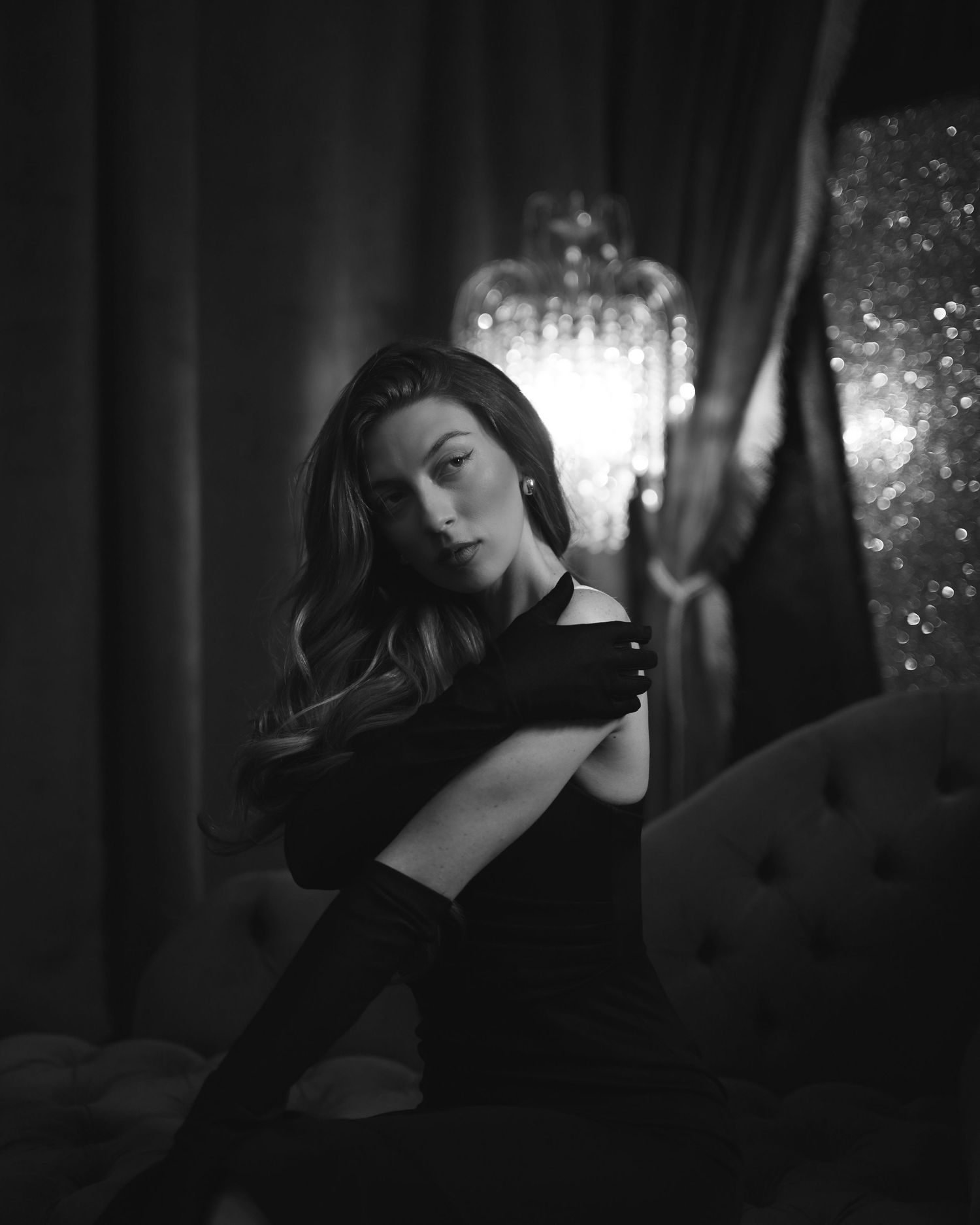Black and white portrait of elegant woman in off-shoulder dress and gloves seated near glowing chandelier.
