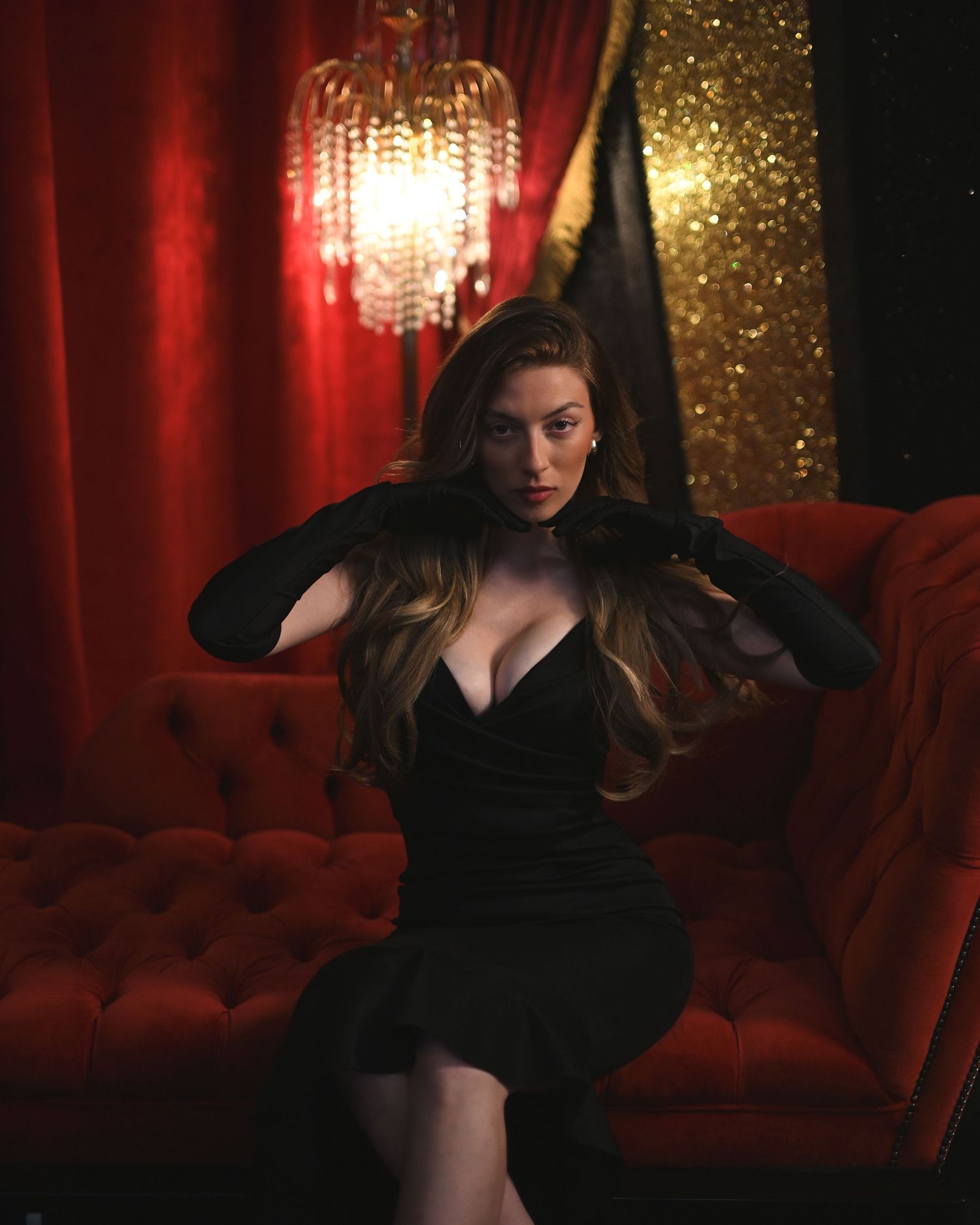 Woman in black dress and gloves poses on red velvet sofa under crystal chandelier in dramatic red room.