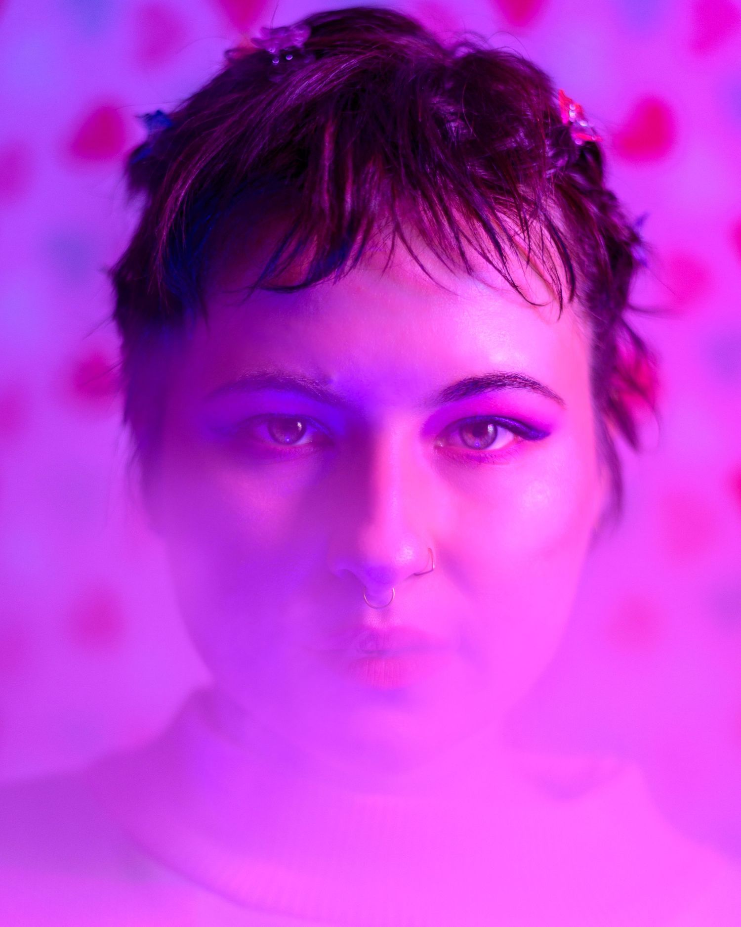 Young non-binary person with short hair and accessories poses in dreamy pink-purple neon light with heart-patterned background.