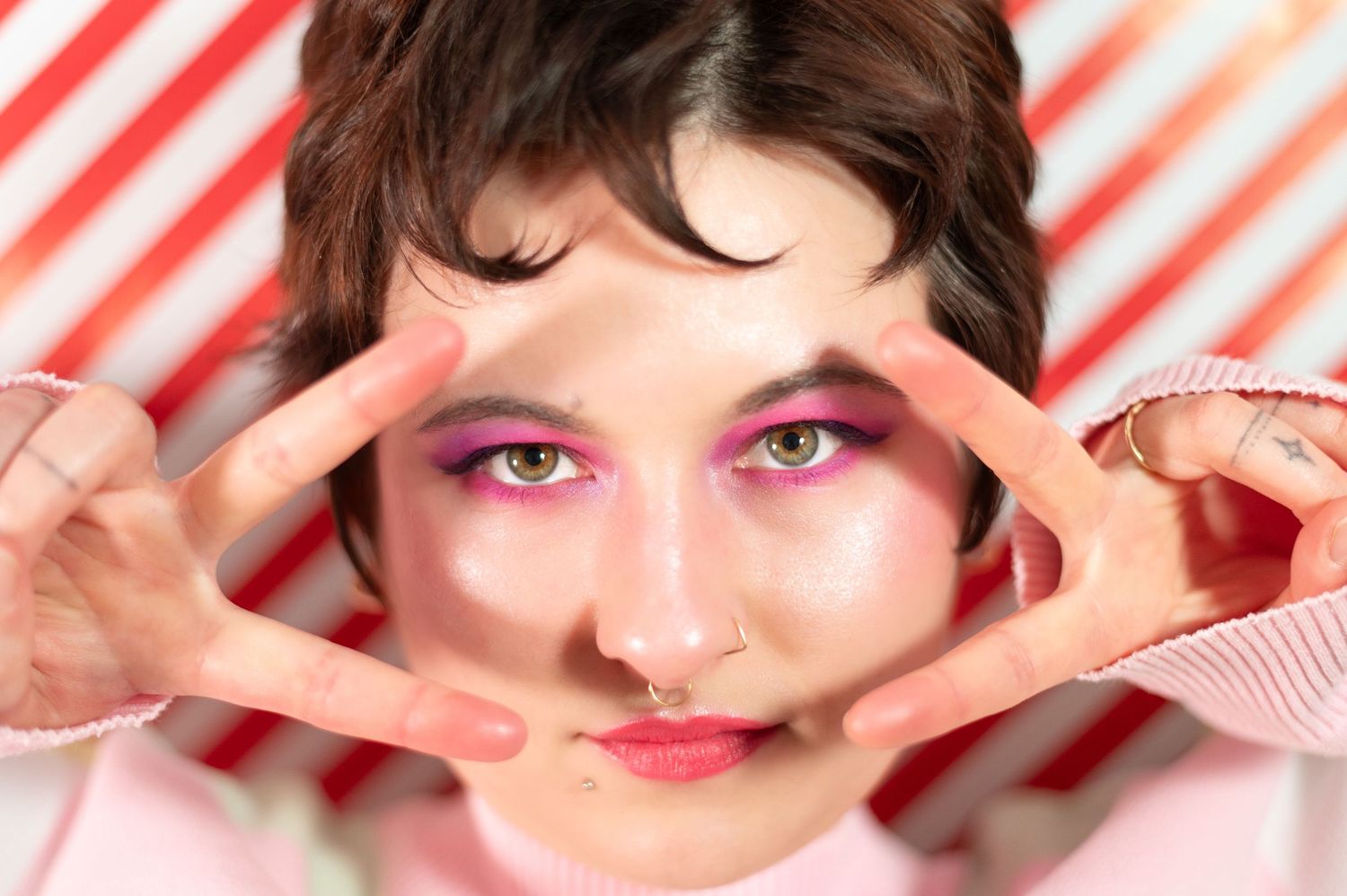 Close-up of a non-binary person with bold pink eye makeup and green eyes, framing their face with their hands against a striped background.