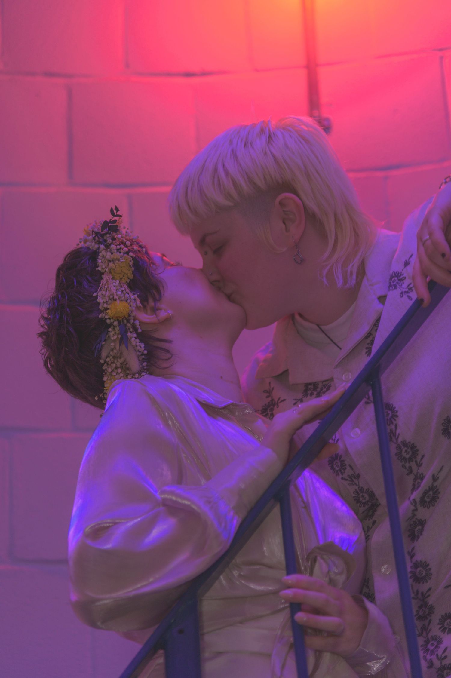 Two people sharing a romantic kiss under pink and purple neon lights, one wearing a floral crown.