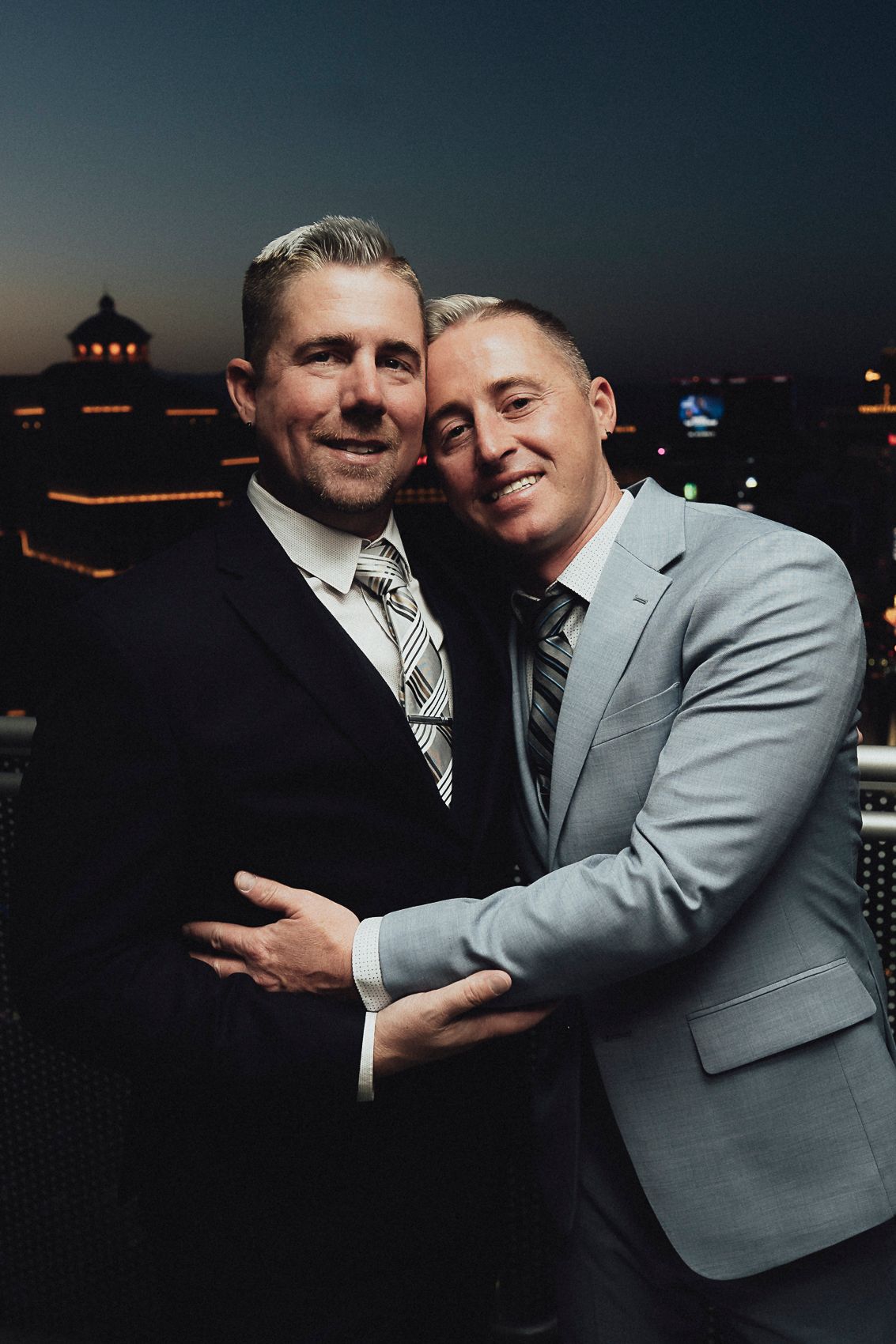 Two grooms in suits embrace romantically on rooftop at night with city lights behind them.