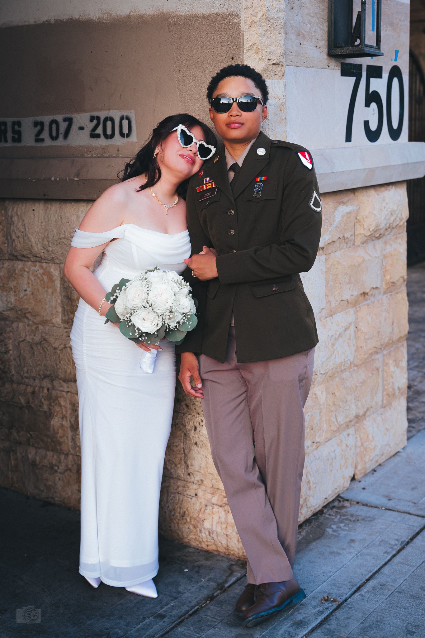 Military groom and bride in white dress pose with sunglasses and bouquet against stone building.