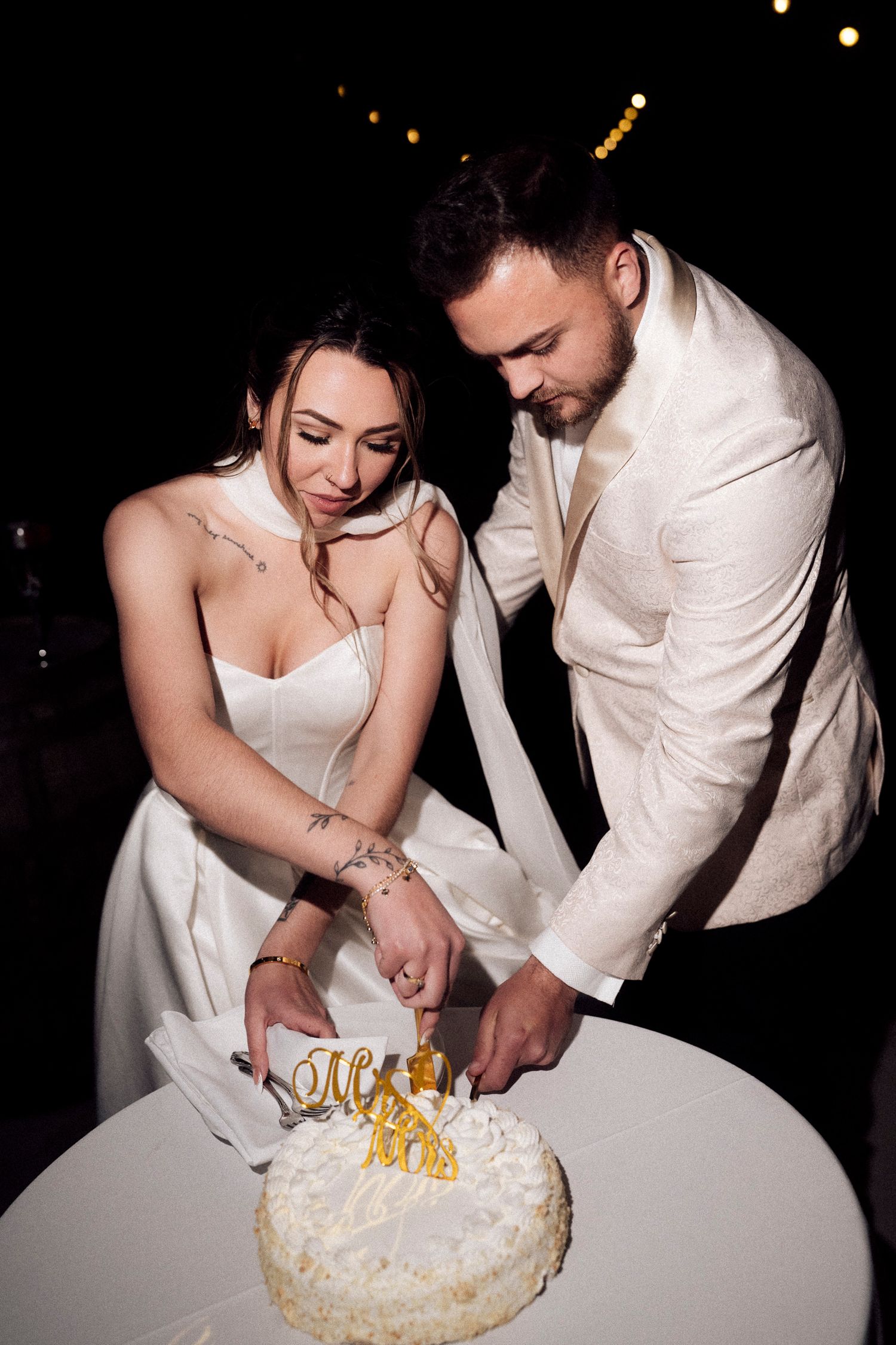 Bride and groom cutting white wedding cake with gold decorations at night celebration.