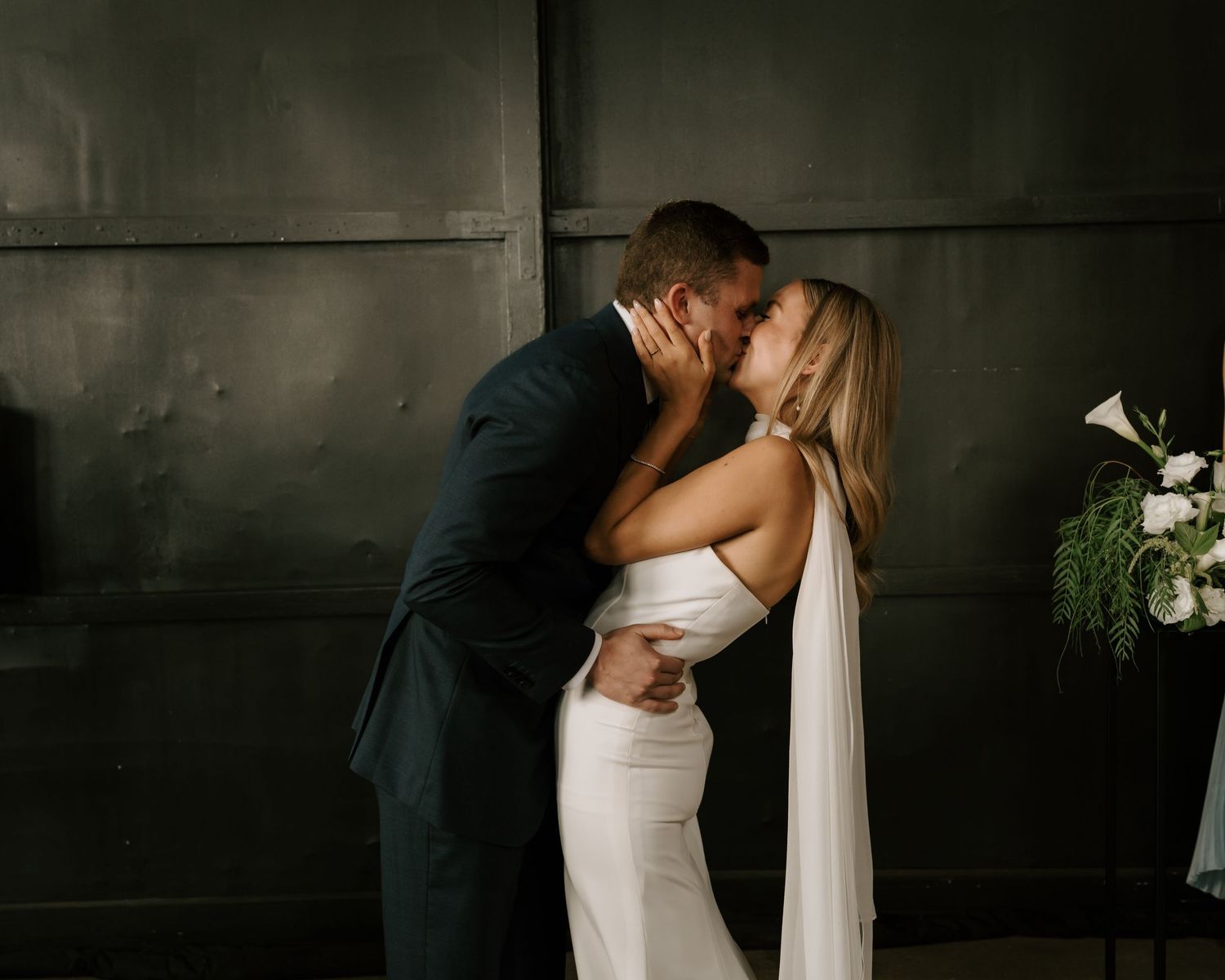 Bride and groom share a romantic first kiss in an elegant dark-walled venue, bride wearing a sleek white gown.