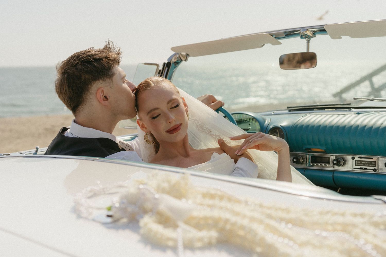 Couple in wedding attire sharing a romantic moment in a vintage teal convertible car on a sunny beach.