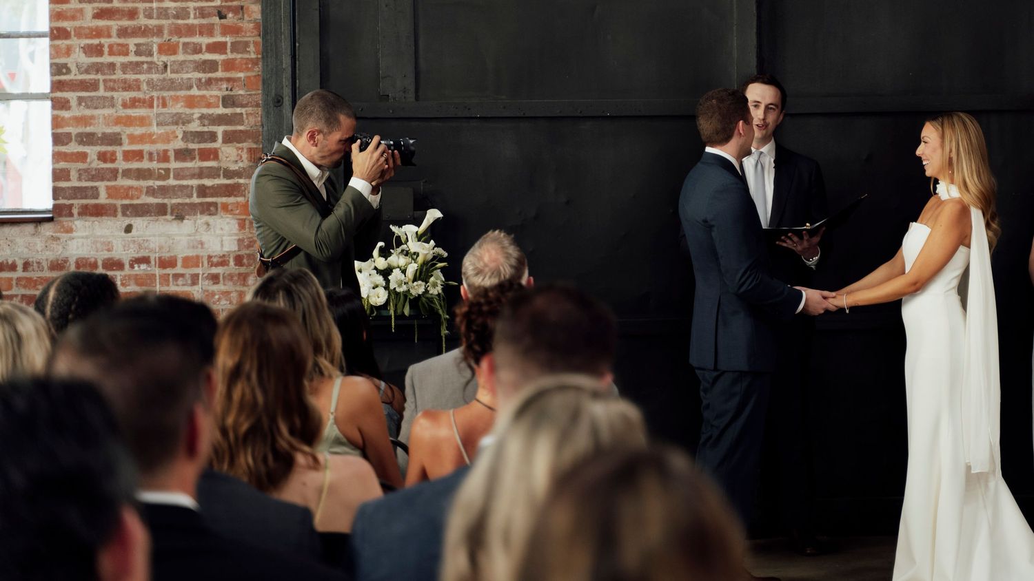 Emotional wedding ceremony moment with guests watching as groom wipes tears during vows indoors.