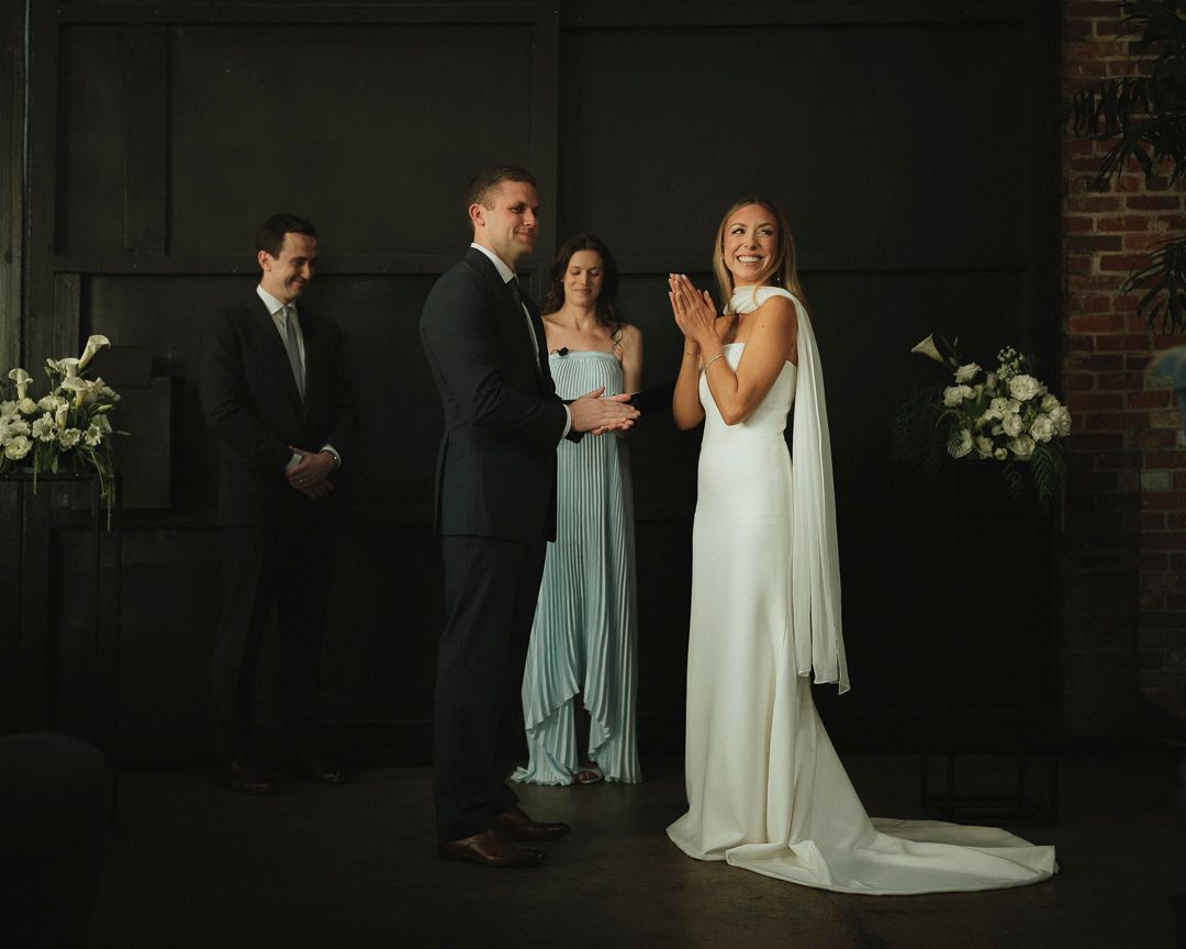 Bride claps joyfully during indoor wedding ceremony while groom holds hands with officiant in sage bridesmaid dress.