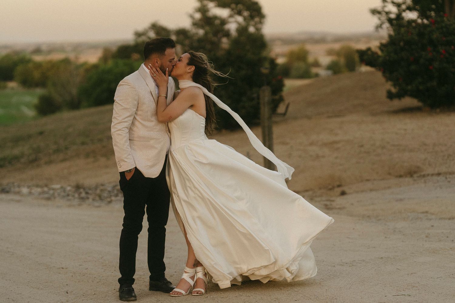 A couple shares a romantic kiss at sunset, the bride's white gown flowing in the wind on a dusty outdoor path.