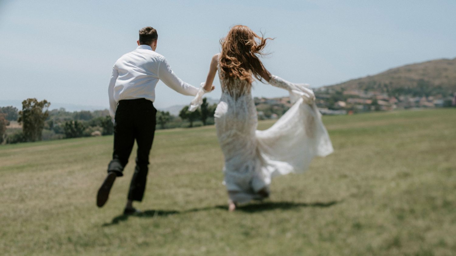 A joyful bride and groom running hand in hand across a lush green field on their wedding day.