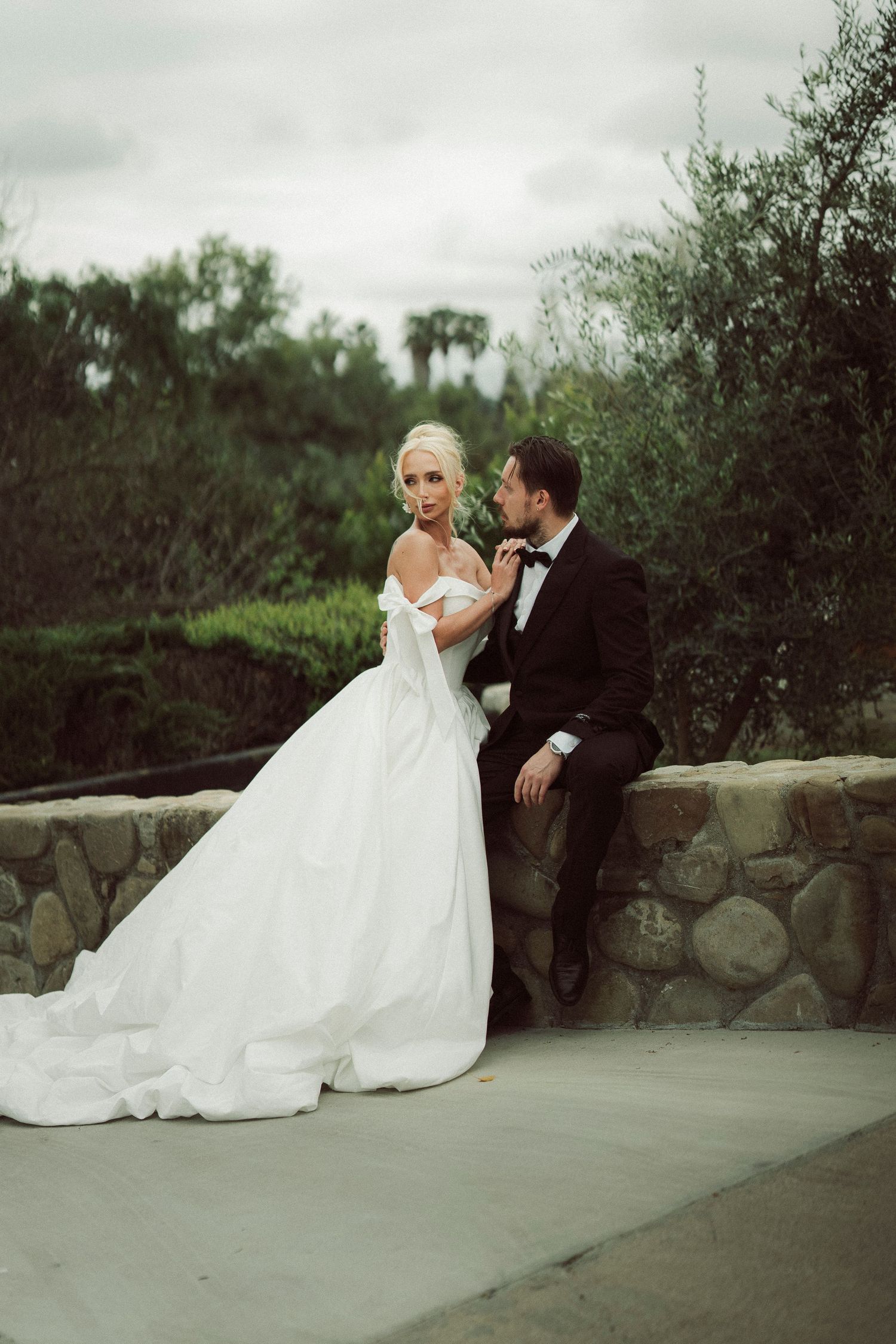 Bride in off-shoulder white gown and groom in tuxedo pose outdoors on stone wall.