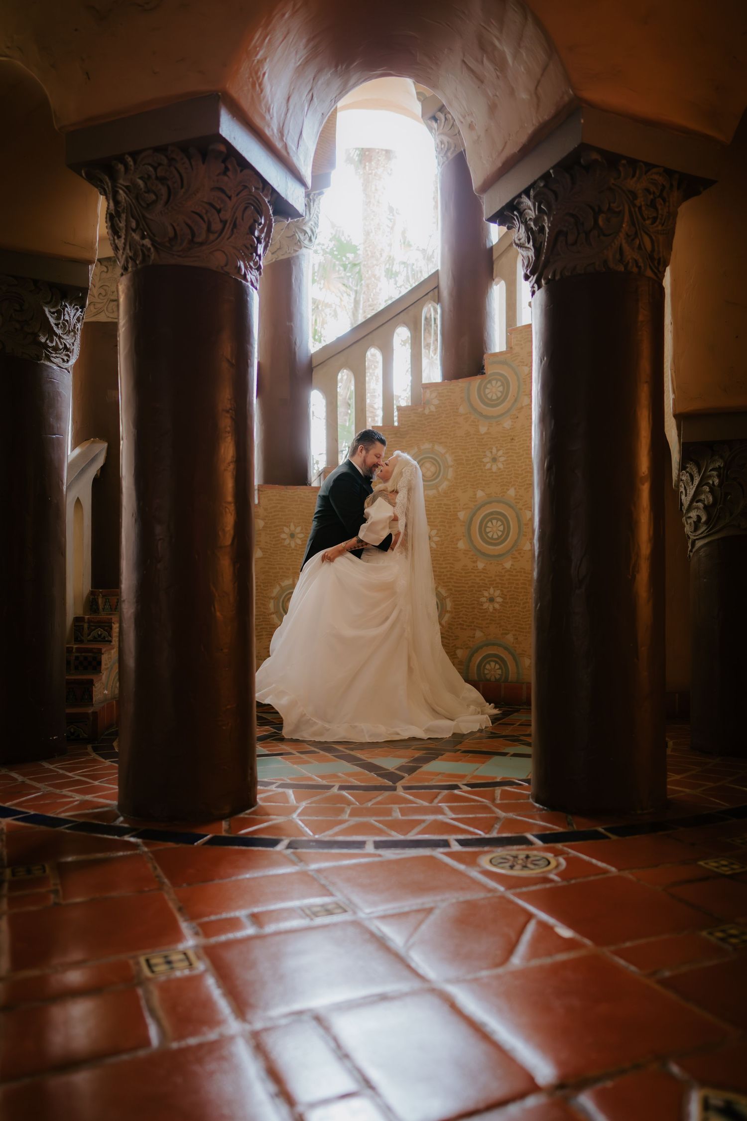 Bride and groom share romantic kiss framed by ornate columns in elegant historic venue.