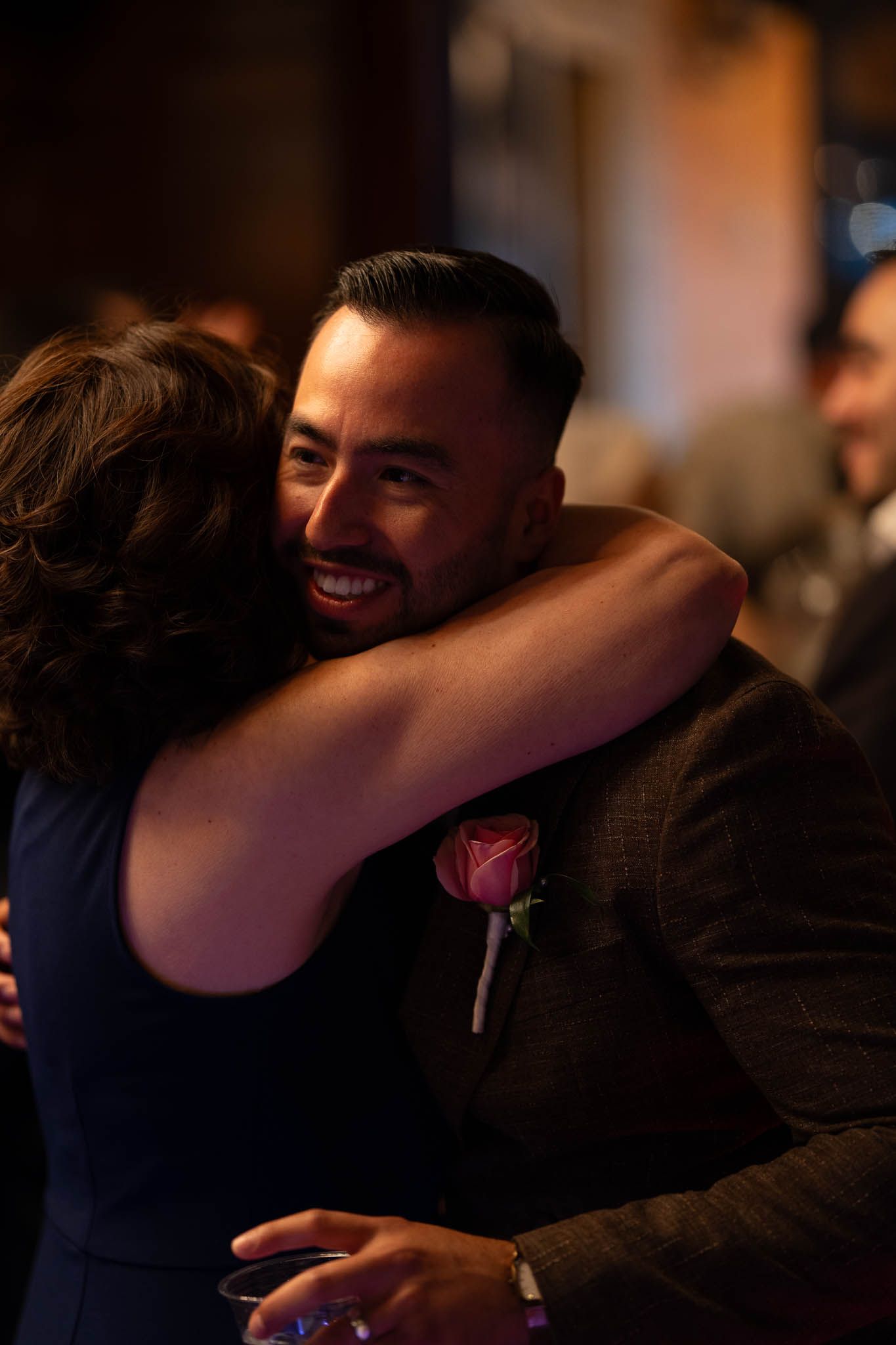 Smiling groom in suit with rose boutonniere shares warm embrace at wedding reception.
