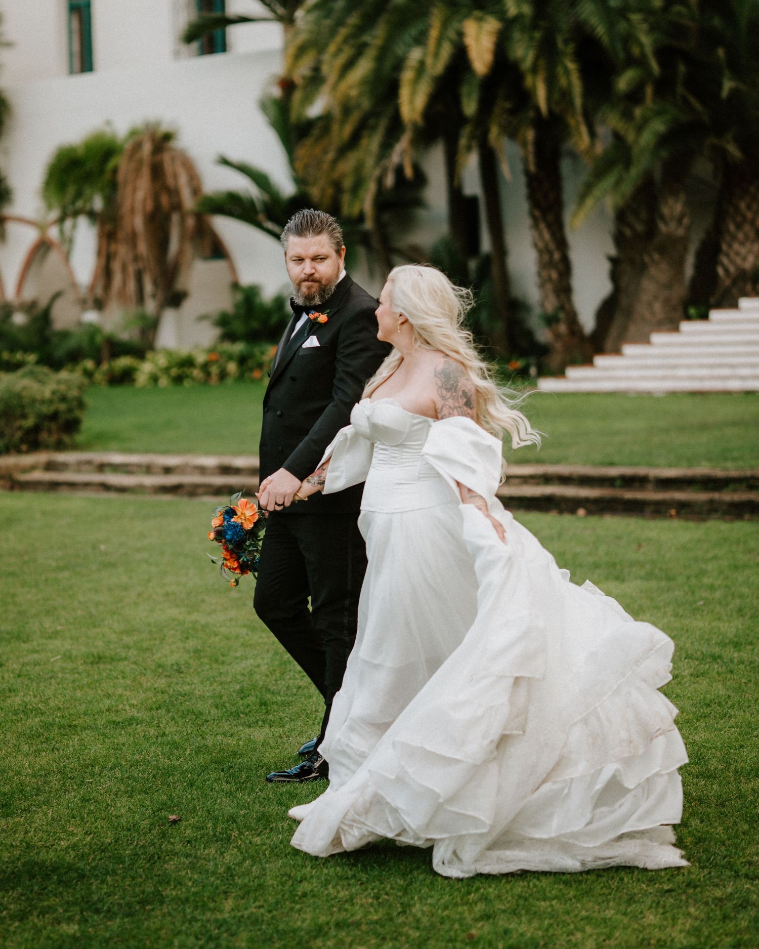 Bride in layered white gown and groom in black tuxedo walk hand in hand on green lawn.