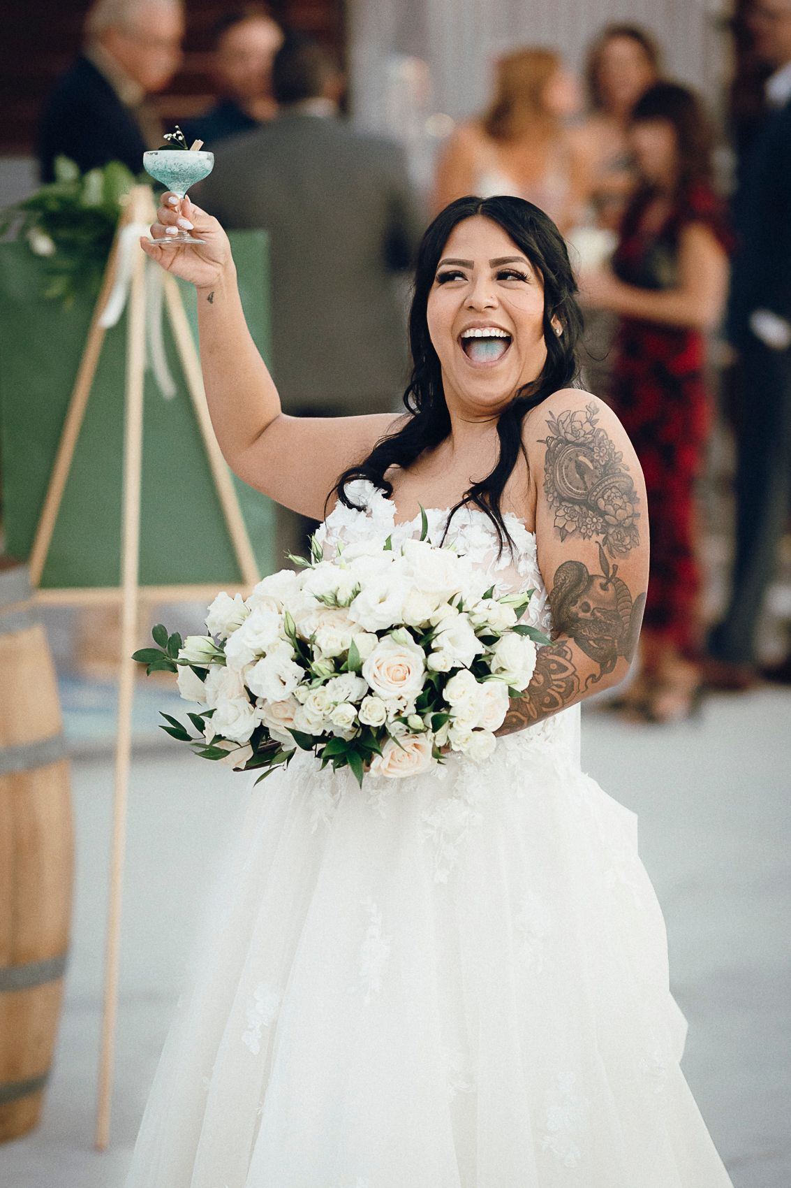 Joyful tattooed bride in white ballgown raises cocktail glass while holding white rose bouquet.