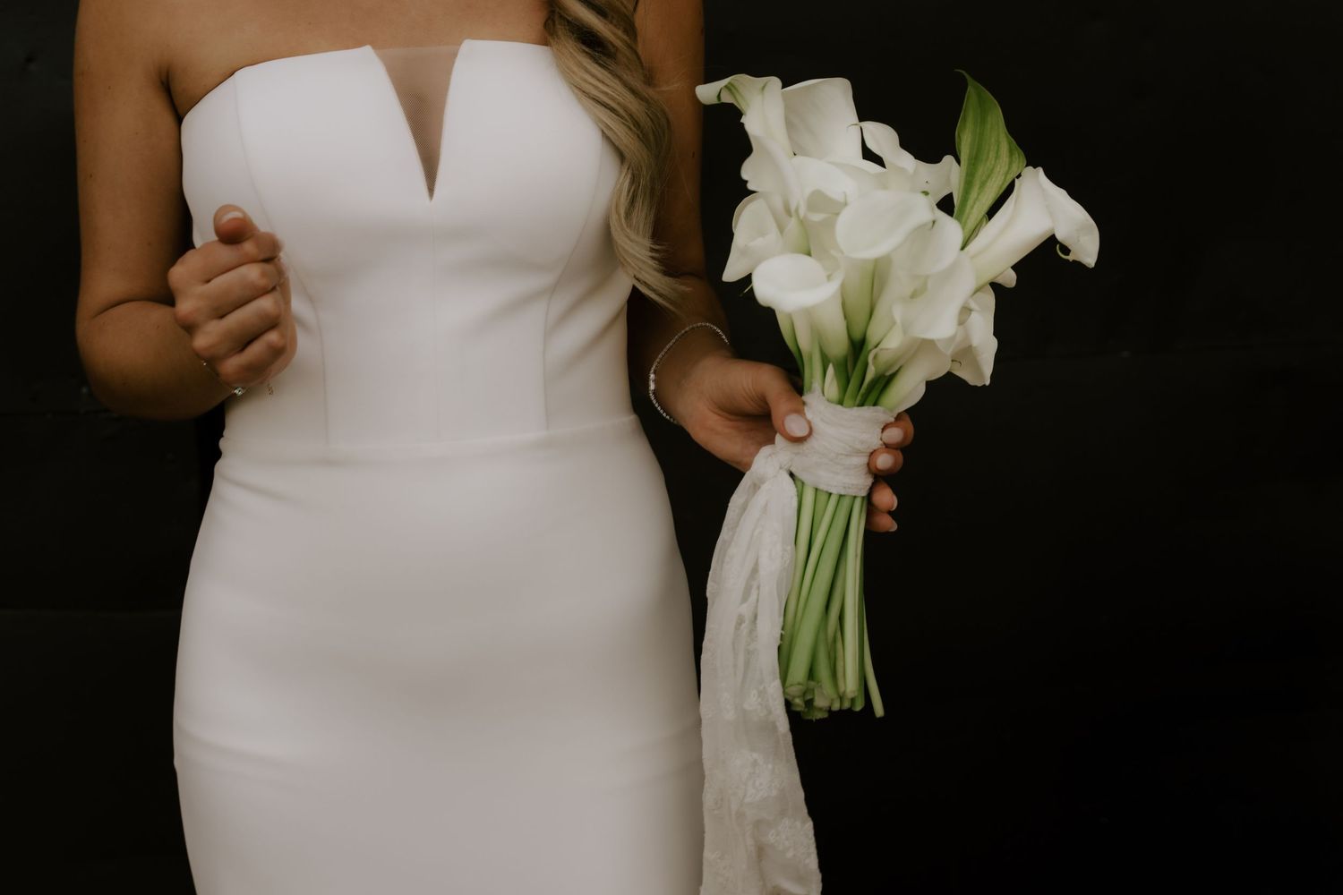 Bride in a sleek white strapless wedding gown holding an elegant bouquet of white calla lilies.