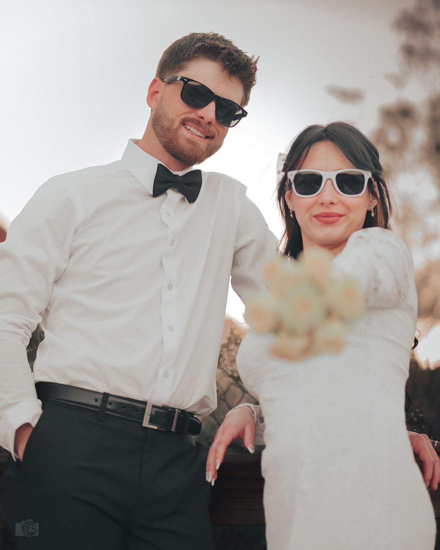 Stylish bride and groom wearing sunglasses pose together with bouquet at outdoor wedding.