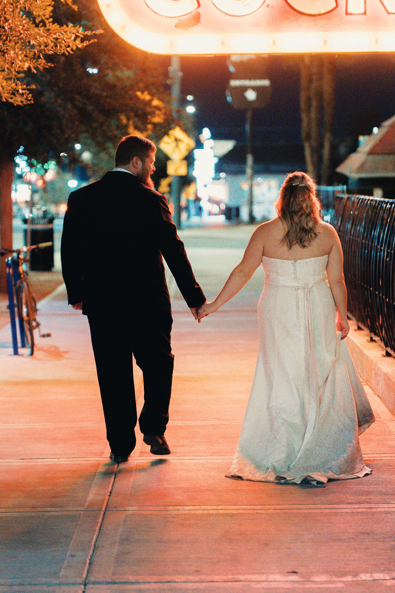 Bride and groom walk hand in hand along city sidewalk under glowing neon lights at night.