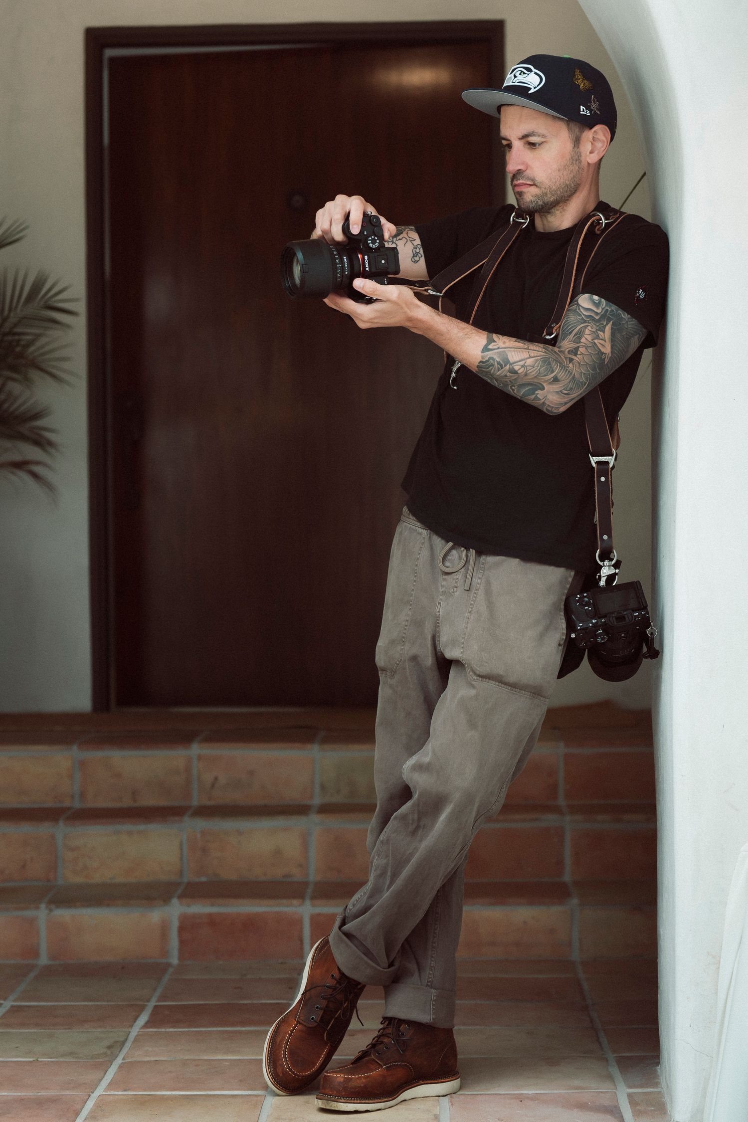 Tattooed male photographer in black tee and cap reviewing photos on DSLR camera while leaning against wall.
