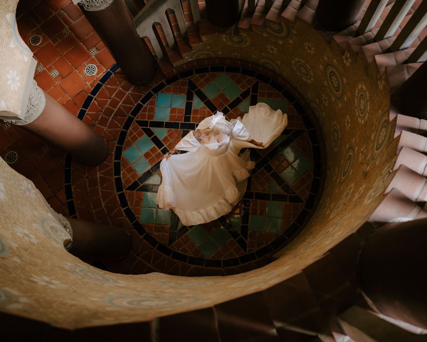 Aerial view of bride in white gown standing on colorful mosaic floor of spiral staircase.
