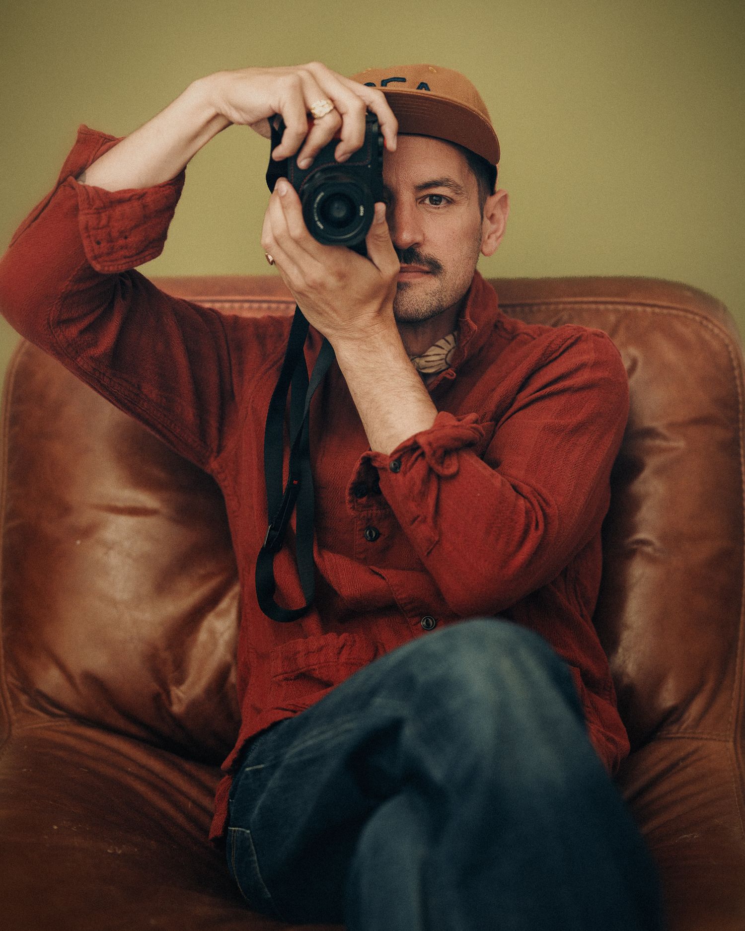 Nick Streeter in red shirt and brown cap sits on leather chair, holding a Leica Q3 camera up to his eye.