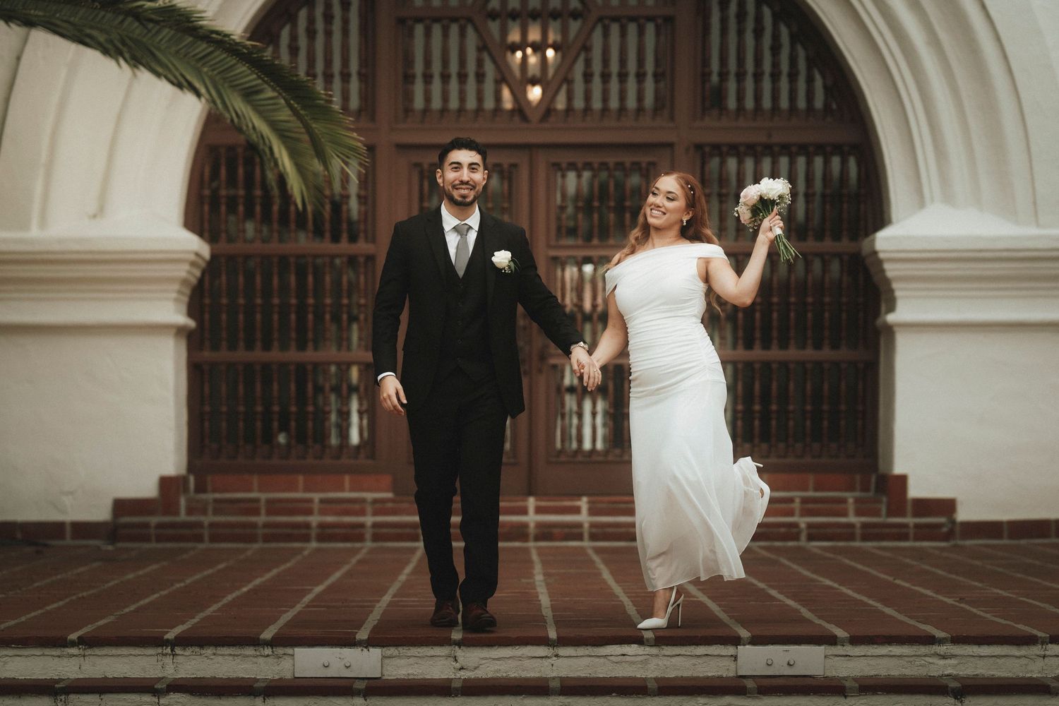 Bride and groom hold hands walking down steps outside a historic building on their wedding day.