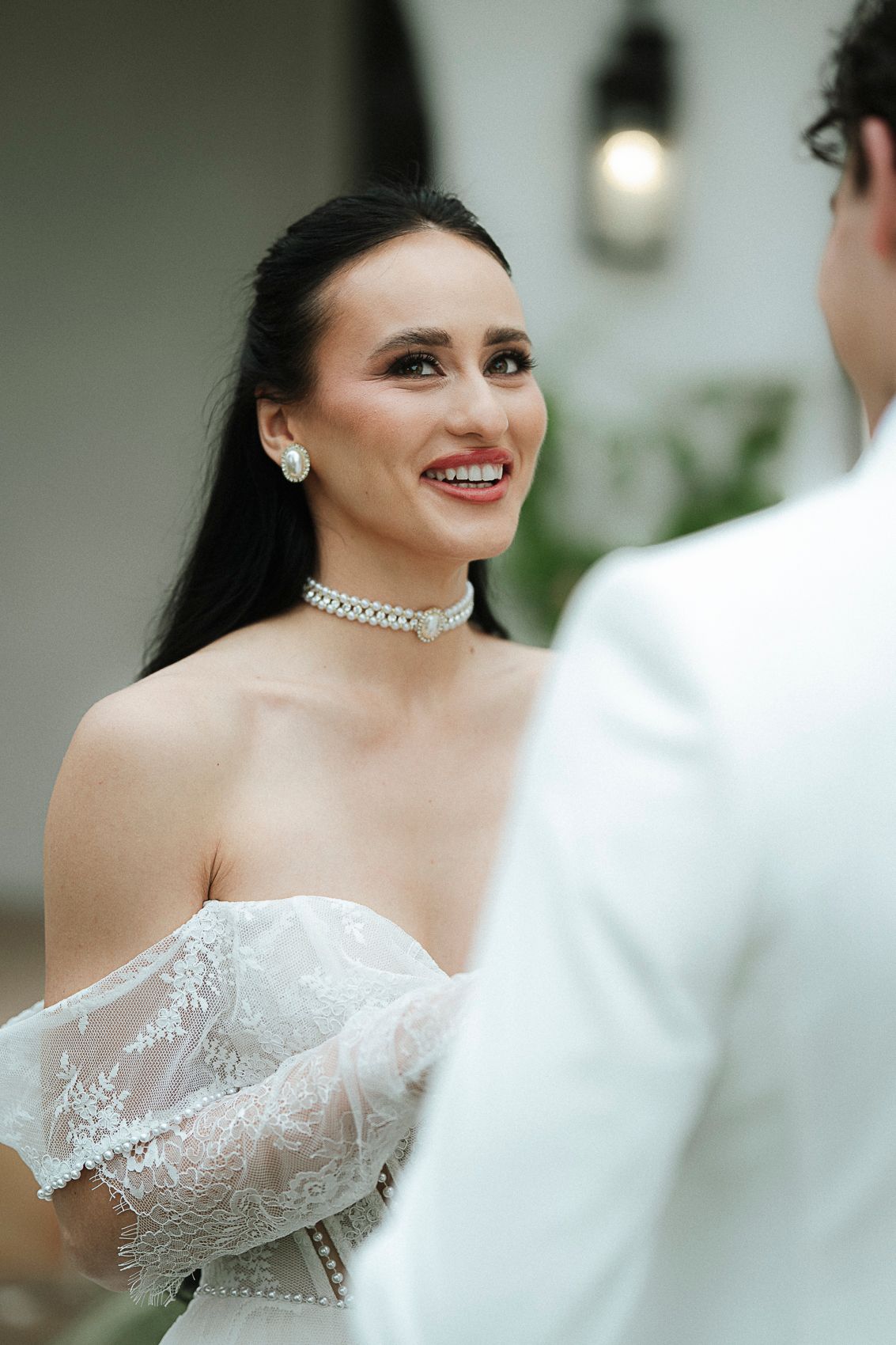 Radiant bride in off-shoulder lace gown smiles wearing diamond choker at outdoor ceremony.
